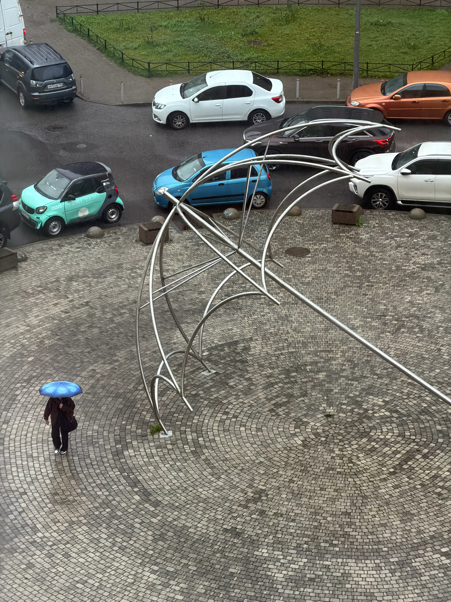 An elevated, high-angle view captures a scene on a rainy day in Saint Petersburg, Russia, focusing on a courtyard with cobblestone paving. In the foreground, a person walks away from the viewer, holding a bright blue umbrella. To their left, a teal and black smart car is parked. In the background, several other cars are visible: a white sedan, a dark-colored SUV, a blue hatchback, and an orange sedan. The cars are parked along a wet asphalt road. A large, abstract metal sculpture, resembling an overturned umbrella, is prominently featured in the mid-ground. The weather appears overcast and damp, contributing to the wet appearance of the road and paving stones. The scene is framed by a grassy area and a black metal fence in the upper portion of the image.