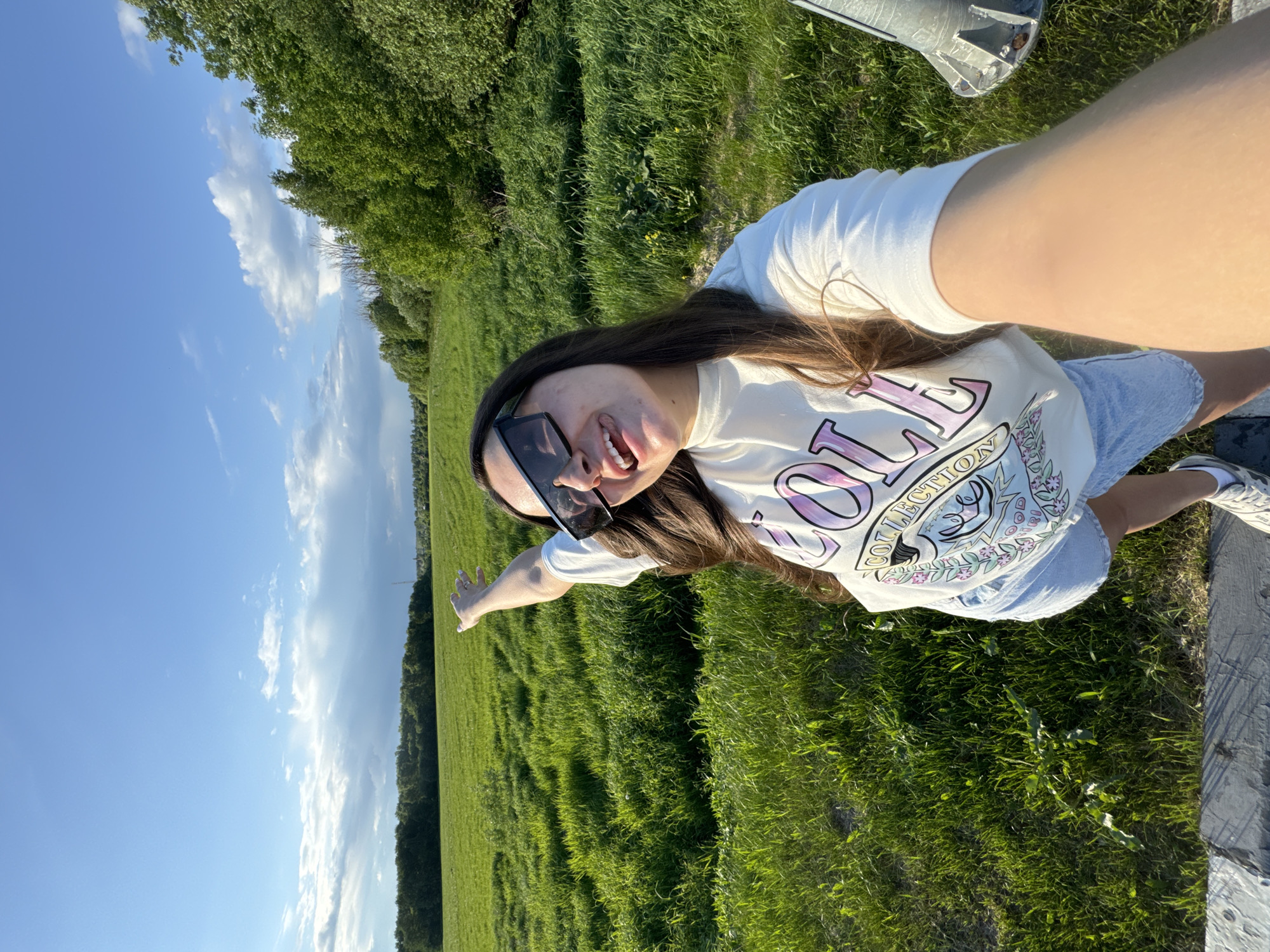 The image shows a young woman in a field, possibly in the area of Bolshiye Vyazyomy, Russia, given the green landscape. She is wearing sunglasses, a white T-shirt with a graphic design, and denim shorts. She is smiling with her tongue showing and pointing with her left arm towards the field. The background includes a grassy field, trees, and a blue sky with clouds. A section of a grey surface can be seen in the bottom right of the image.