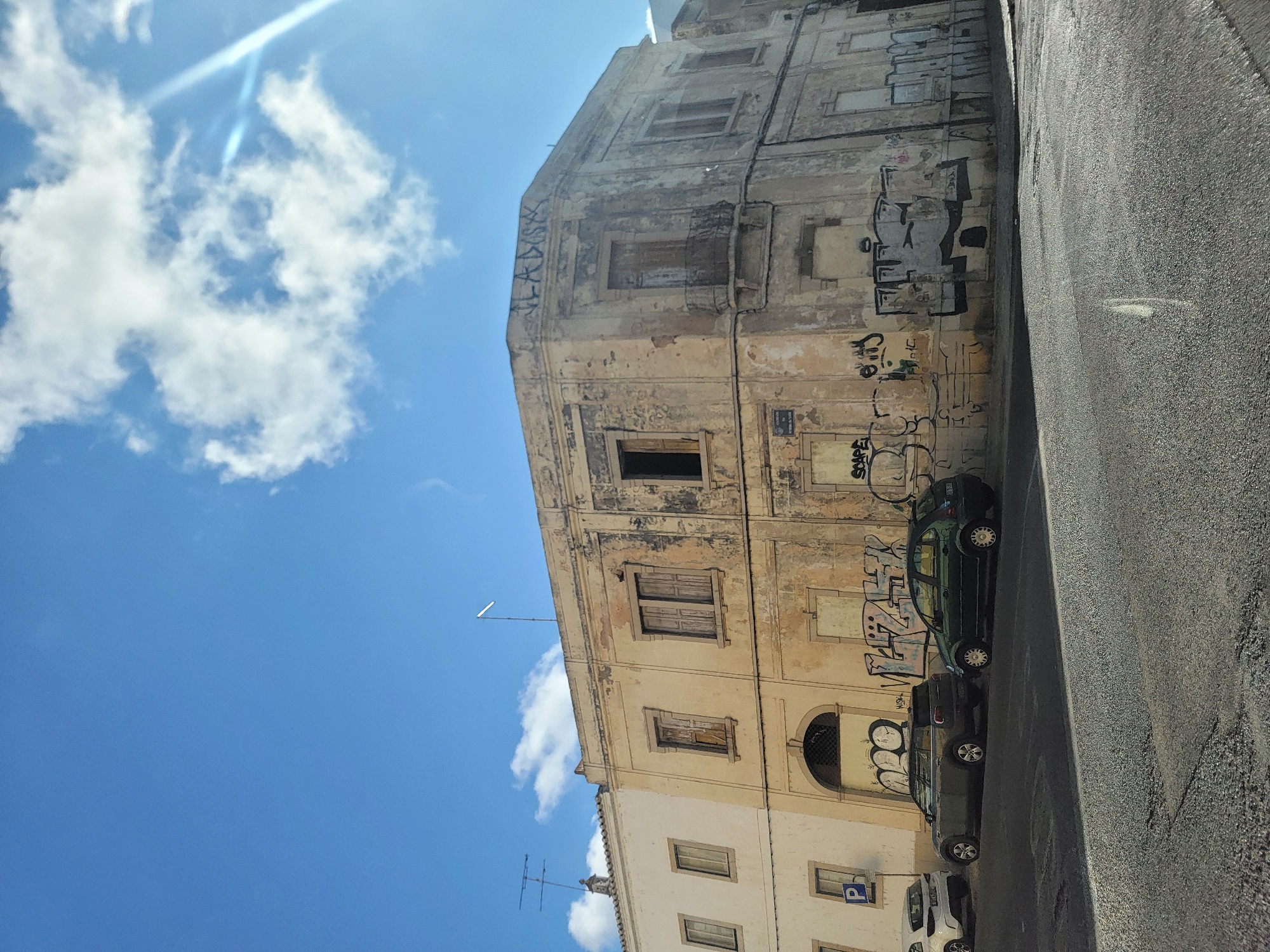 The image shows an old, weathered building in Faro, Portugal, with extensive graffiti on its facade. The building appears to be on a street corner, with a road and several parked cars in the foreground. The sky is blue with scattered clouds, and sunlight is casting a lens flare on the right side of the image. The building's architecture suggests it is historic, with boarded-up windows and peeling paint. The graffiti is in various styles and colors, covering a significant portion of the lower walls. Three cars are visible: a white hatchback, a dark grey station wagon, and a dark green hatchback. The street in front of the building is paved, and a street sign with a blue "P" for parking is visible on the left. The overall impression is one of urban decay and street art in a sunny, Mediterranean environment.