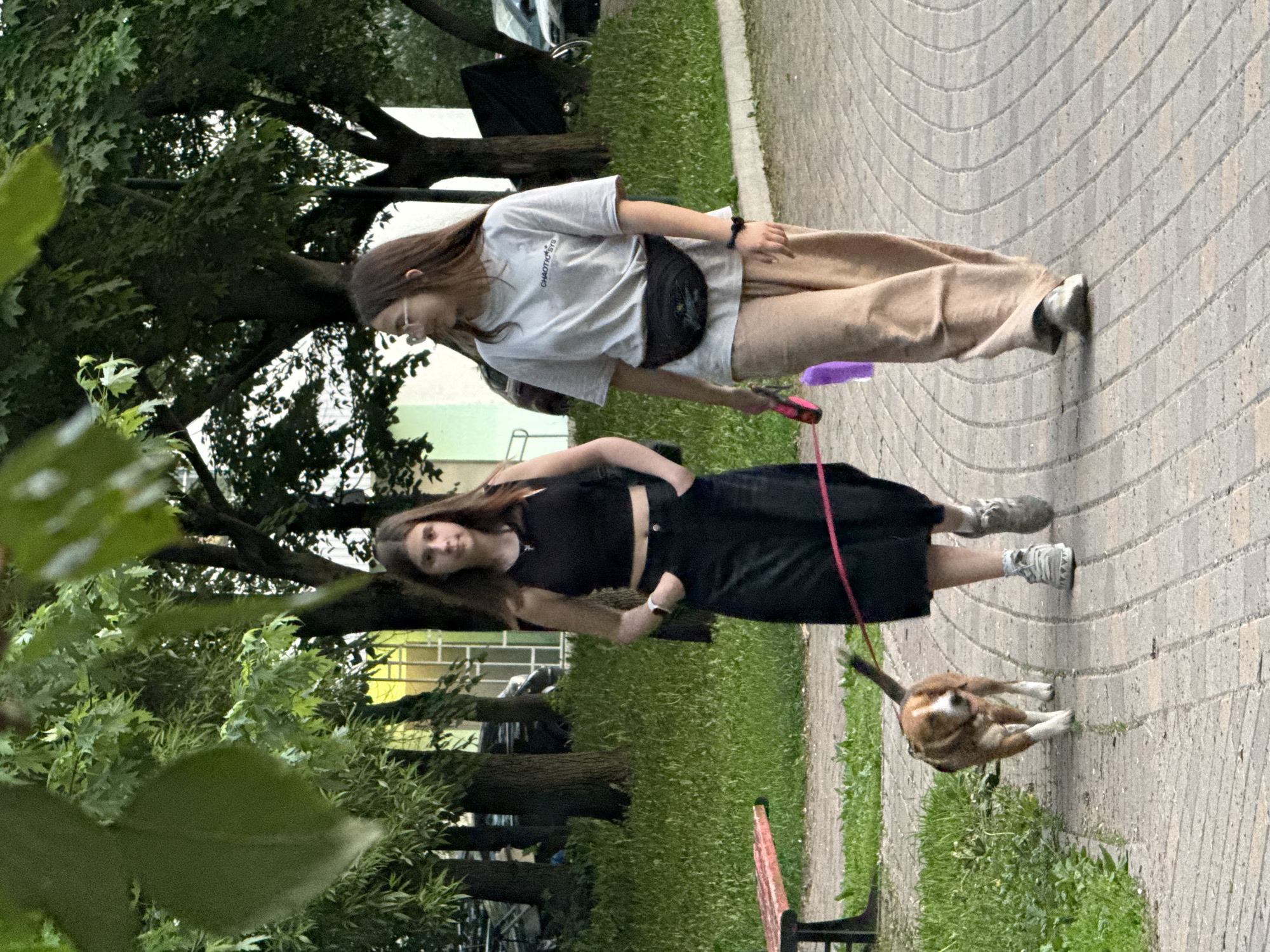 Two young women are standing on a paved pathway, one holding a leash connected to a small dog. The woman closer to the foreground is wearing a black crop top and a long black skirt, while the other woman is wearing a light-colored t-shirt and beige pants. They are in an outdoor setting with trees and greenery visible in the background. A bench is partially visible in the lower right corner. The location is Moscow, Russia.