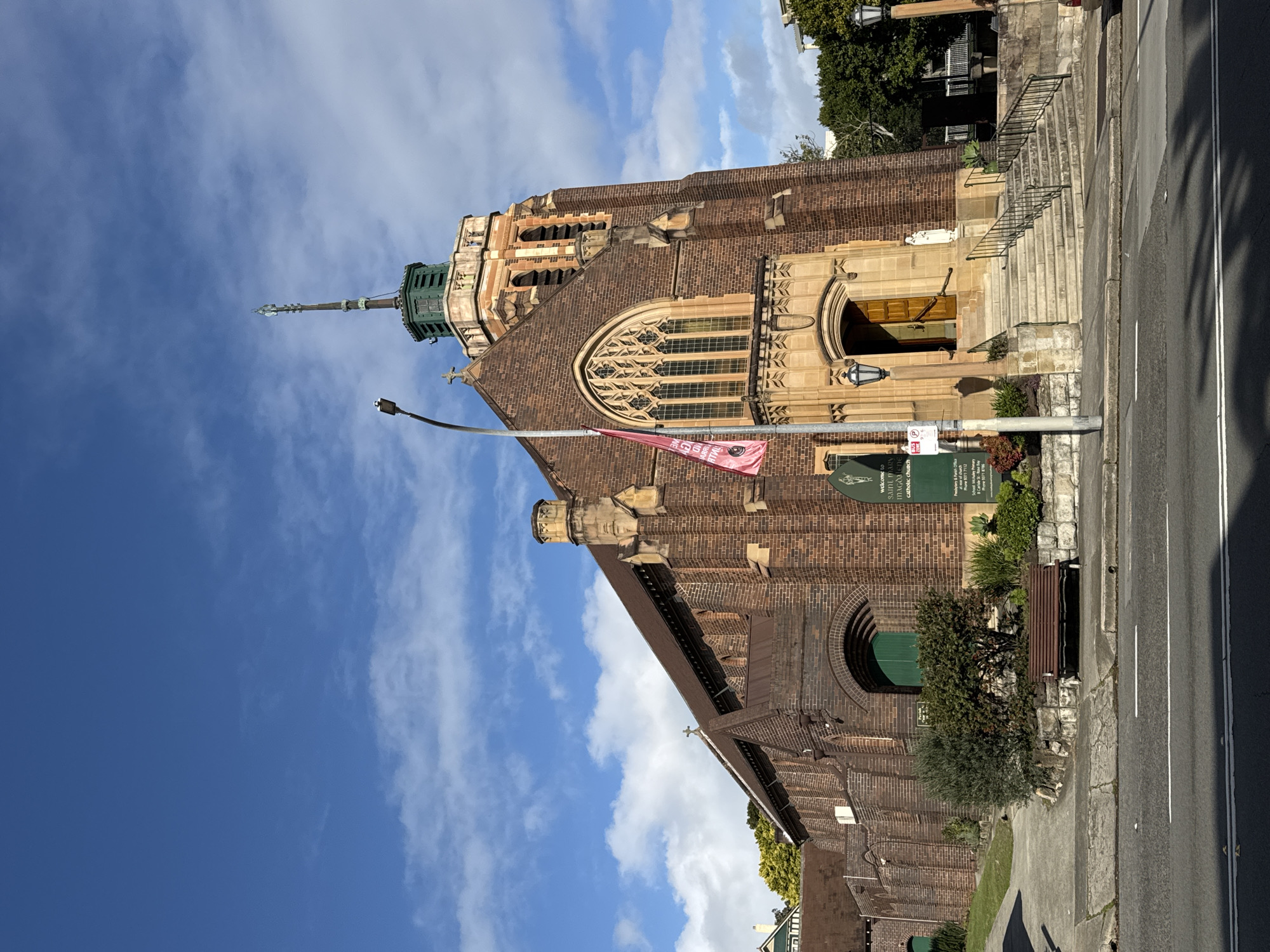 The image shows the exterior of St. Mary Magdalene Catholic Church in Vaucluse, Australia, on a sunny day with a blue sky and scattered clouds. The church is constructed of red brick with sandstone accents and features a prominent tower with a green spire. The main entrance is up a set of stone steps, with a wooden door. A pink banner is hanging from a flagpole on the left side of the entrance. To the left of the church, a paved path leads up to a grassy slope. A park bench is situated on the path. A street runs in front of the church, with a traffic light and a signpost. Green trees and bushes are visible surrounding the church and on the right side of the image.