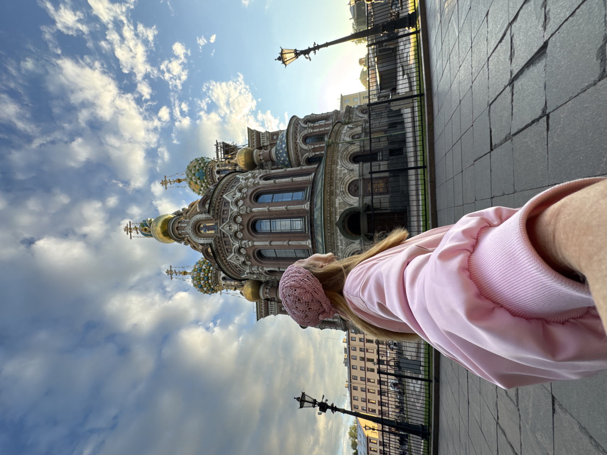 This media file is a photograph taken outdoors in Saint Petersburg, Russia. The primary subject is a person, seen from behind and from the shoulder up, wearing a pink knitted hat and a light pink jacket. Their blonde hair is visible. They are looking towards a large, ornate building with multiple domes, some of which are golden and others are decorated with colorful patterns. This building is likely a church, given its architectural style. The background shows a clear blue sky with fluffy white clouds, suggesting it is daytime, possibly late afternoon or early evening due to the warm light on the building. A lamppost is visible in front of the building, and a black metal fence separates the foreground from the background. The ground in the foreground is paved with large, dark gray square tiles. The overall scene conveys a sense of tourism or sightseeing. There is no visible text in the image.
