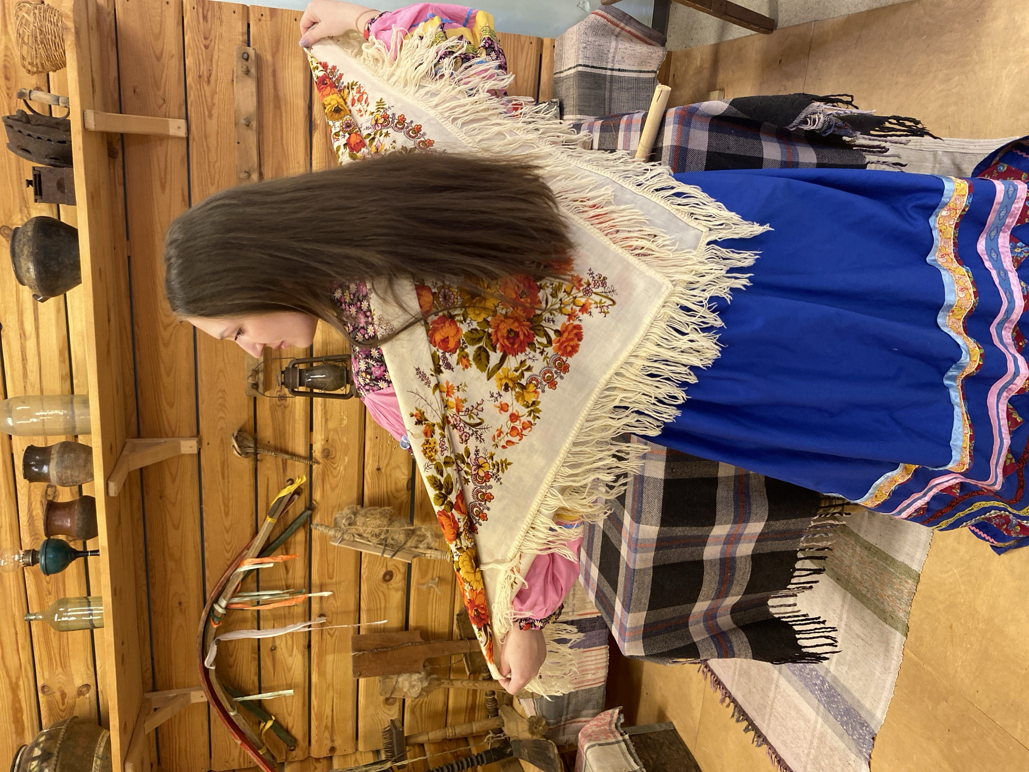 A young woman is depicted in profile, positioned indoors against a wooden plank wall in what appears to be a cultural exhibit or museum setting in Markovskii, Russia. She has long, dark brown hair that falls over her left shoulder. She is dressed in traditional attire, consisting of a pink long-sleeved blouse with a subtle floral pattern, and a vibrant blue full skirt adorned with a colorful, patterned trim featuring stripes and ribbons in pink, yellow, and red.
Draped over her shoulders is a large, cream-colored shawl, intricately patterned with vivid orange, red, and yellow floral motifs and green foliage. The shawl is edged with a long, cream-colored fringe, which she holds with both hands as if adjusting it or posing.
The background features wooden shelves displaying various rustic and antique items. On the left, these include old cast-iron irons, a dark metal pot, several glass bottles or vases, and a small woven basket. An old metal lantern is visible further to the right. Behind the woman, several plaid blankets or woven textiles in dark and light patterns are visible, contributing to the historical or folk ambiance of the scene. No text is visible in the image.