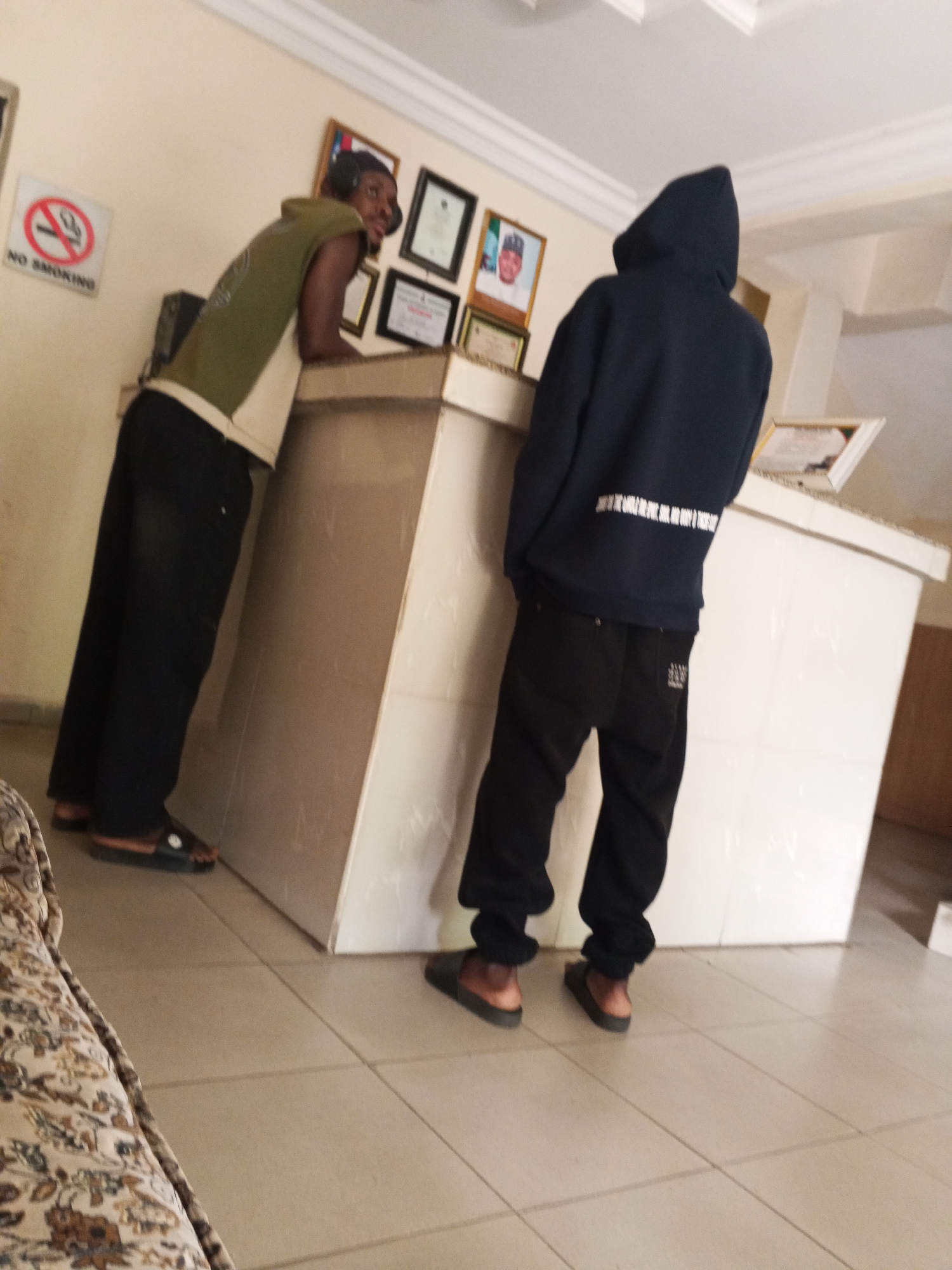Two individuals stand near a reception desk inside a building. The person on the left, facing the reception desk, is a Black male, wearing a green sleeveless top, black trousers, and sandals. He is leaning on the desk with his arms. To his right is a Black person wearing a dark-blue hoodie, black trousers, and sandals. The hoodie is pulled over their head. They are standing next to the desk. The reception desk is white. On the wall behind the desk are several framed pictures and a "No Smoking" sign. The floor is tiled. A section of floral-patterned furniture is visible in the lower-left corner. The location is Unknown, Unknown.