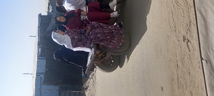 This image, captured in Khan Yunis, Palestinian Territory, depicts a daylight outdoor scene where several individuals are seated on a two-wheeled cart on an unpaved road. The cart, constructed with a wooden or metal frame and dark, worn tires, transports at least four people.
From left to right:
1. A person largely obscured, wearing dark, full-body attire (possibly an abaya or niqab), is seated on the far left of the cart.
2. Next to them, a woman wears a white hijab and a long, dark red/purple patterned dress. Her hands are clasped in front of her.
3. To her right, another woman wears a black hijab and a maroon tracksuit. She holds a white cup or container in her right hand, and a white bag is visible on her lap.
4. Partially visible on the far right is an individual wearing a white shirt.
The ground beneath the cart is an unpaved, dusty, and sandy surface, suggesting a dirt road, with a clearer, possibly paved or more compacted area in the immediate foreground. In the background, several temporary structures are visible, constructed from corrugated metal sheets, tarpaulins, and other makeshift materials, indicative of an informal settlement or camp. The sky above is clear and light blue, and the scene is brightly illuminated by direct sunlight, casting shadows from the cart and individuals.