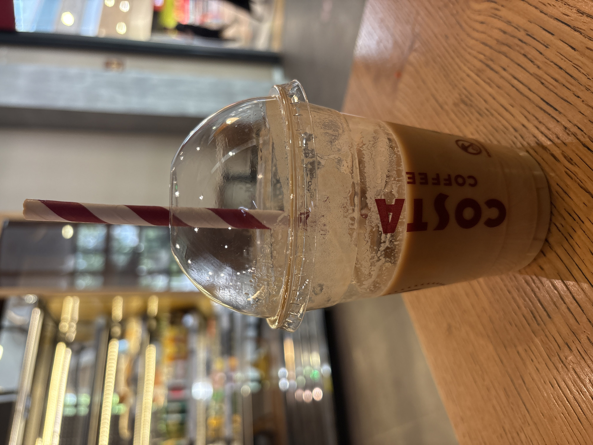 The image shows a nearly empty plastic cup with a clear domed lid, resting on a wooden surface. A red and white striped straw extends from the lid into the cup. The cup has condensation marks on the inside and some remaining light brown liquid. The logo on the cup clearly reads "COSTA COFFEE" in bold, dark red lettering. The background is blurred but appears to be an indoor setting with display shelves and soft lighting, possibly a coffee shop or cafe. The image is taken from a slightly elevated angle, focusing on the cup and straw. The lighting suggests it could be daytime, but the interior setting makes it difficult to determine definitively. There are no people visible in the frame. The overall impression is of a finished beverage, with the cup left on a table.