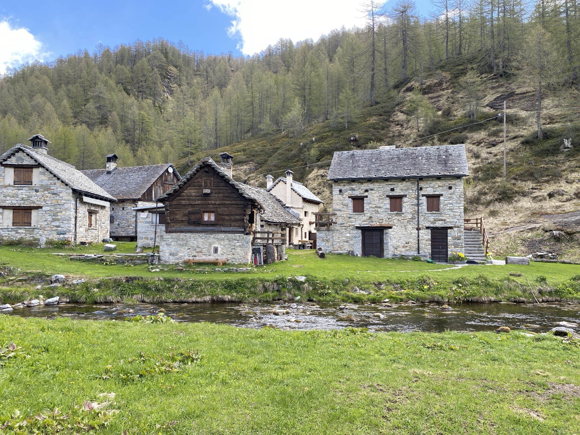 The image captures a picturesque scene in Baceno, Italy, showcasing a cluster of traditional stone and wood buildings set against a backdrop of a forested hillside under a partly cloudy blue sky. A shallow, clear stream flows in the foreground, bordered by lush green grass.
The architecture features rustic houses with stone walls, slate roofs, and wooden accents. Some structures have visible chimneys. The houses are arranged somewhat haphazardly along the stream bank. A set of wooden stairs ascends the slope next to one of the stone buildings.
The hillside behind the houses is covered with deciduous trees, predominantly in early spring foliage, with some bare branches still visible. The ground is also covered with grass. The overall impression is of a tranquil, rural village nestled within a natural, hilly environment.