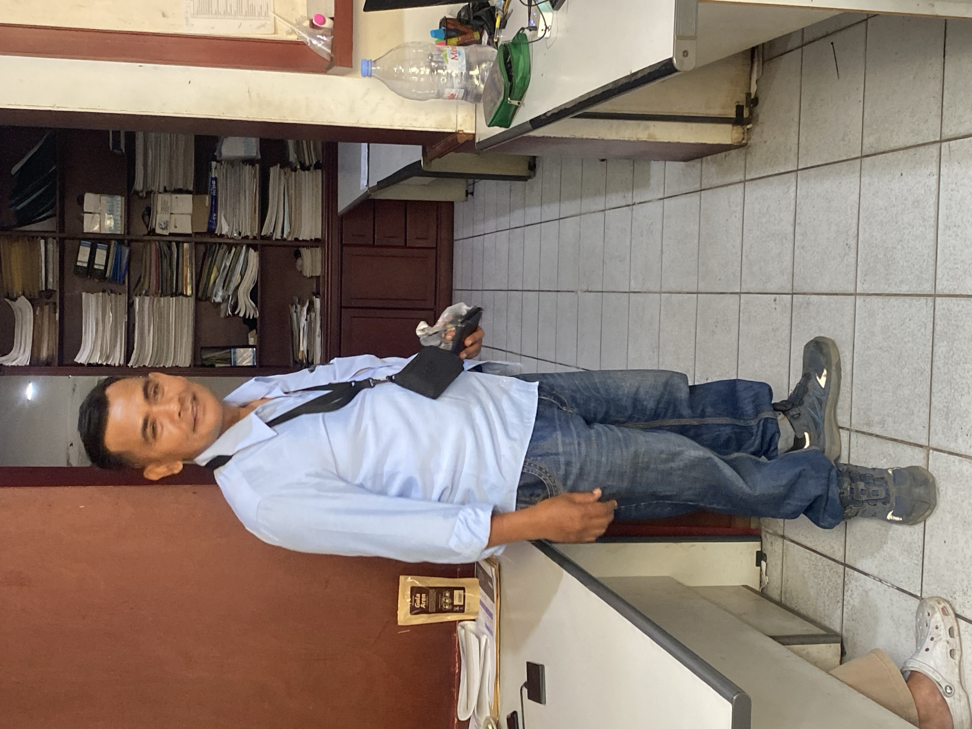 The image depicts a man standing in an office environment in North Jakarta, Indonesia. He is wearing a light blue long-sleeved shirt, blue denim jeans, and dark colored work boots. A black crossbody bag is slung over his shoulder, and he is holding a small dark object in his right hand. The man is standing in front of a large wooden bookshelf filled with stacks of papers and folders. To his left, there is a white desk with some papers and a small black electronic device. To his right, another white desk is visible with a large plastic water bottle, a green pouch, and other office supplies. The floor is tiled with light-colored square tiles. The overall impression is of a functional office space.