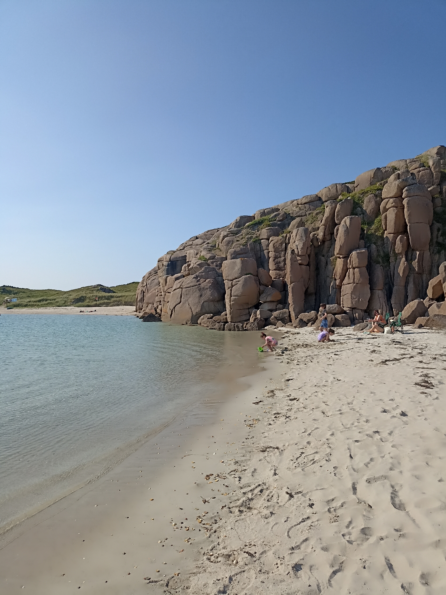 The image depicts a sunny day at a beach in Carnboy, Ireland. A calm, clear body of water meets a sandy shore, with a large, rocky cliff face dominating the right side of the frame. In the distance, grassy dunes are visible. On the sandy beach, a small group of people are present. Three children are playing near the water's edge, one with a green bucket. Further up the beach, two adults are seated in chairs, one appearing to be eating, and a green bag or cooler is beside them. The overall scene is one of a relaxed beach outing.