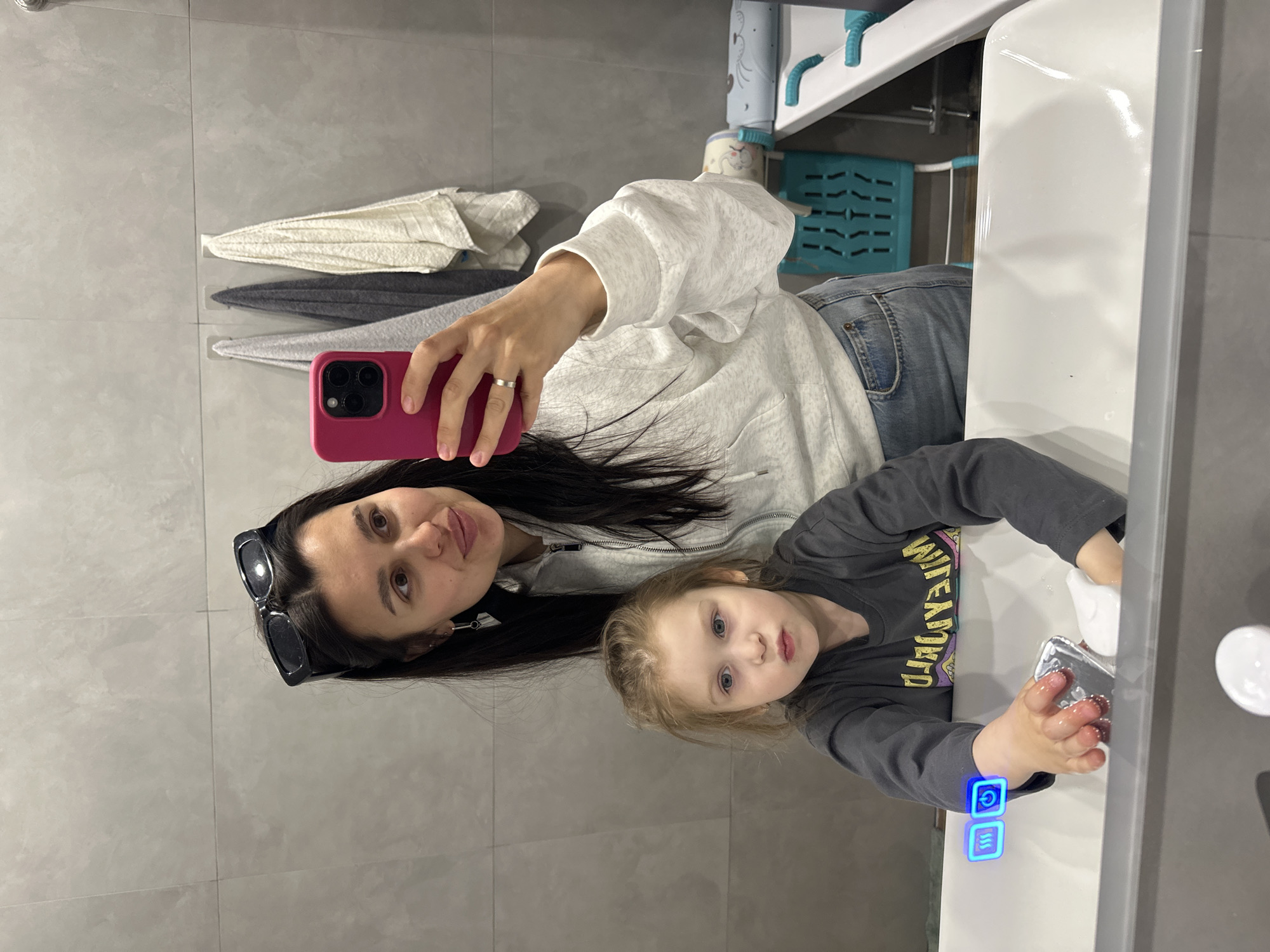 The image depicts a woman and a young child in a bathroom setting. The woman, wearing sunglasses perched on her head and a light-colored hoodie, is taking a selfie with a pink smartphone. The child, wearing a gray long-sleeved shirt, is at the sink, appearing to be washing their hands. The bathroom has gray tiled walls and a white sink. Towels are hung on the wall. Based on the user's prompt of "Chaikovsky, Russia," it can be inferred that this is the location of the scene.