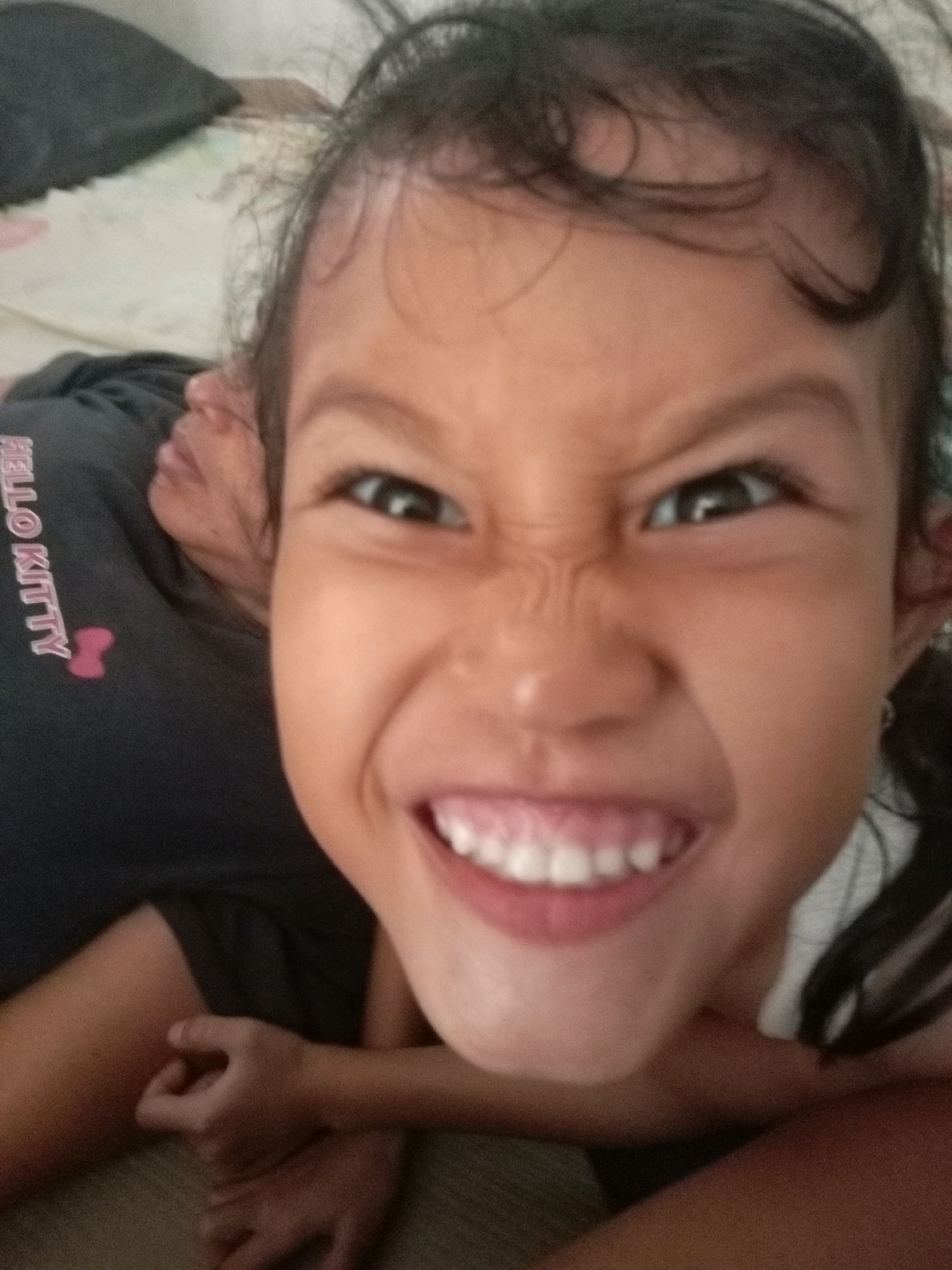 The image shows a close-up portrait of a young girl in Batam, Indonesia. She has dark hair and is making a grimacing facial expression, with her eyes narrowed and her mouth open, revealing her teeth. She is wearing a dark gray t-shirt with a pink "Hello Kitty" logo on the left chest area. The background appears to be a bed or soft surface. The lighting suggests an indoor setting.