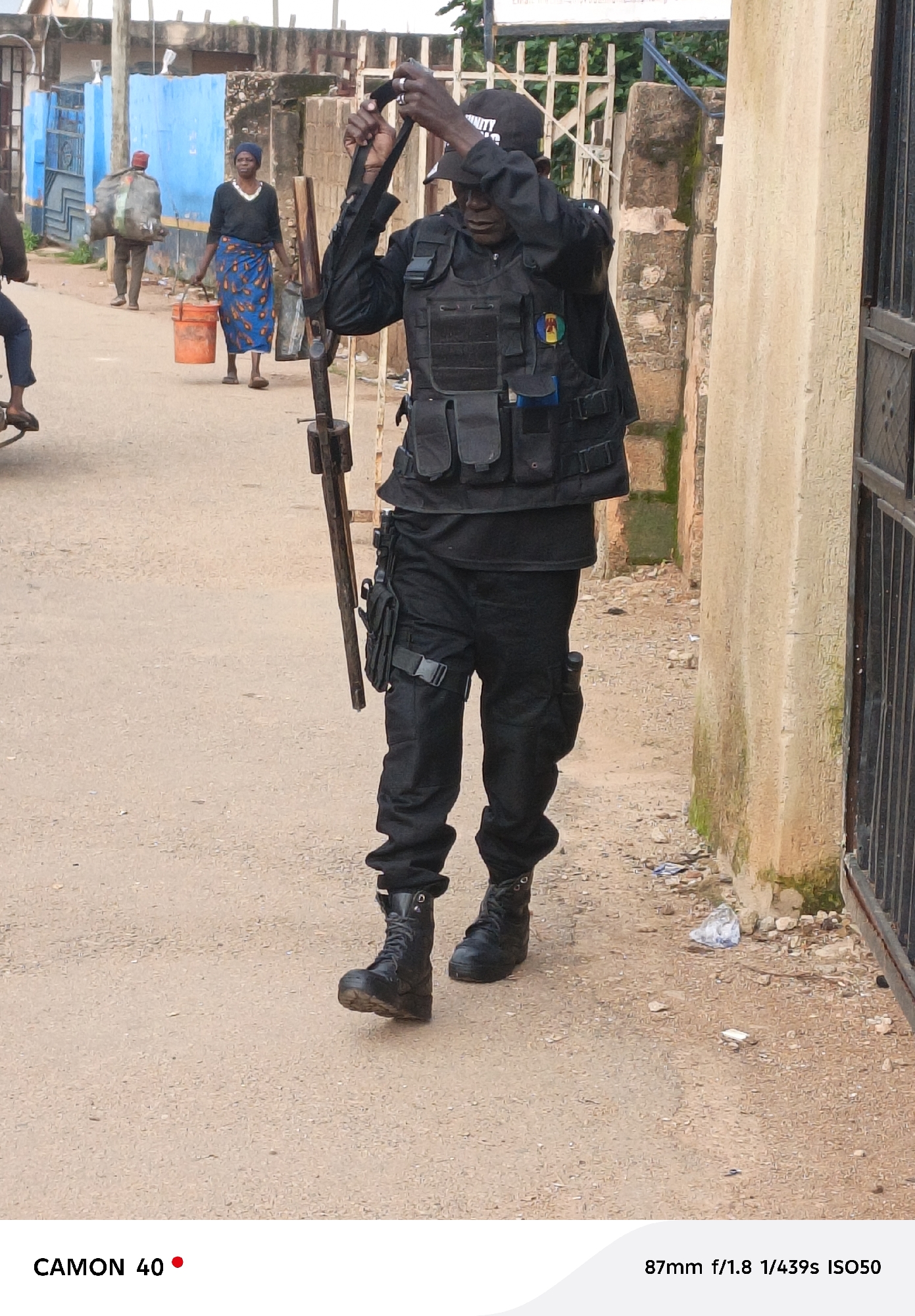 A security guard, wearing a black uniform, tactical vest, and cap with "SECURITY" visible, stands on a dirt road in Jos, Nigeria. He is holding a long, dark object, possibly a baton or rifle, and appears to be taking a photograph with his cell phone. To his left, a woman in a blue and patterned skirt and a dark top carries two buckets, one orange and one grey. Behind her, another person walks away, carrying a large, gray sack over their shoulder. The environment is an urban setting with buildings, a fence, and a gate visible.