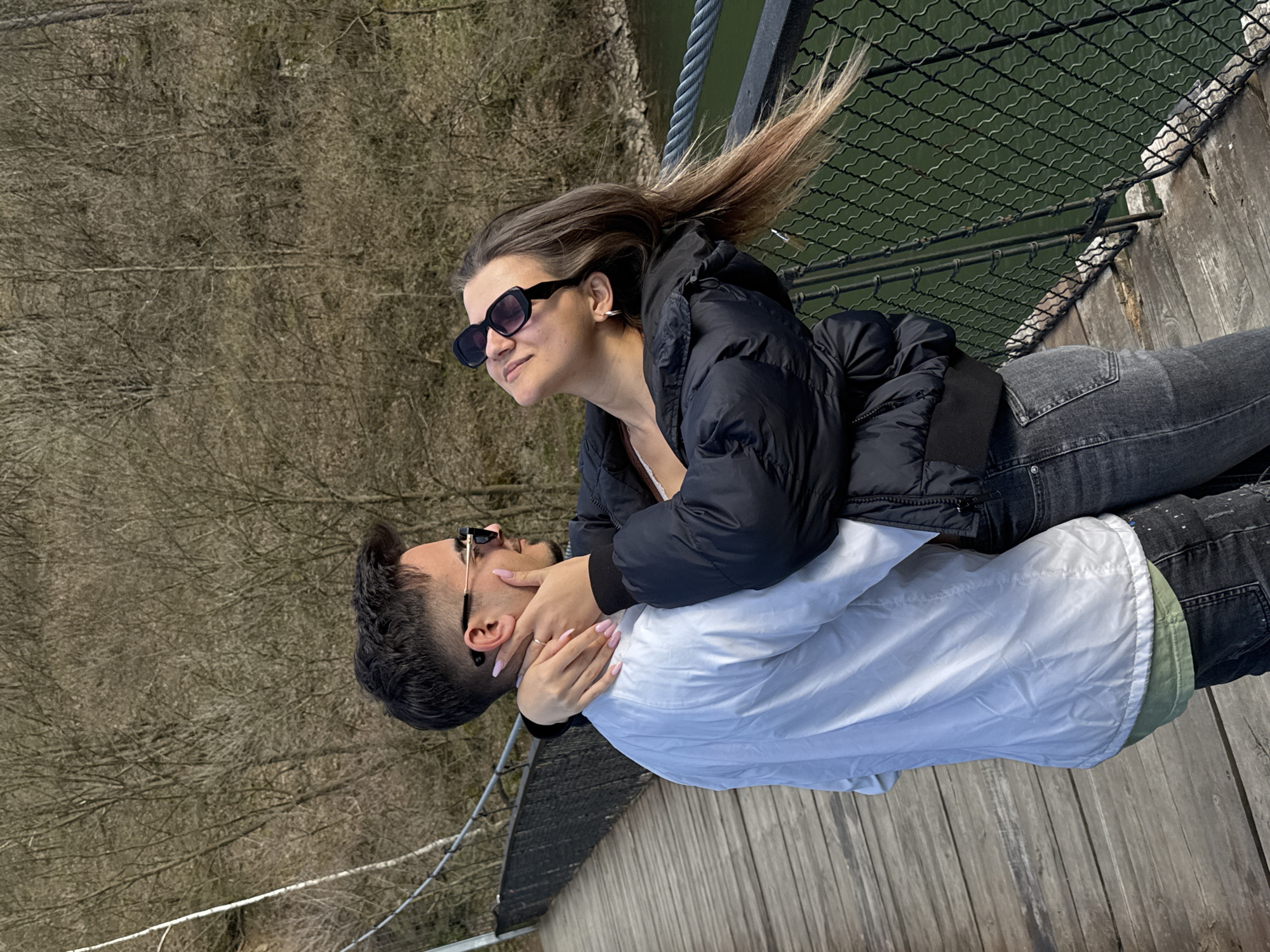 In Dolni Pasarel, Bulgaria, a woman wearing a black puffer jacket and sunglasses leans over a man with sunglasses, holding his face, as they stand on a wooden platform with a wire mesh fence. Behind them is a blurred view of trees without leaves and a dark green body of water. The man is wearing a white jacket over a green shirt. The platform appears to be part of a bridge or elevated structure.