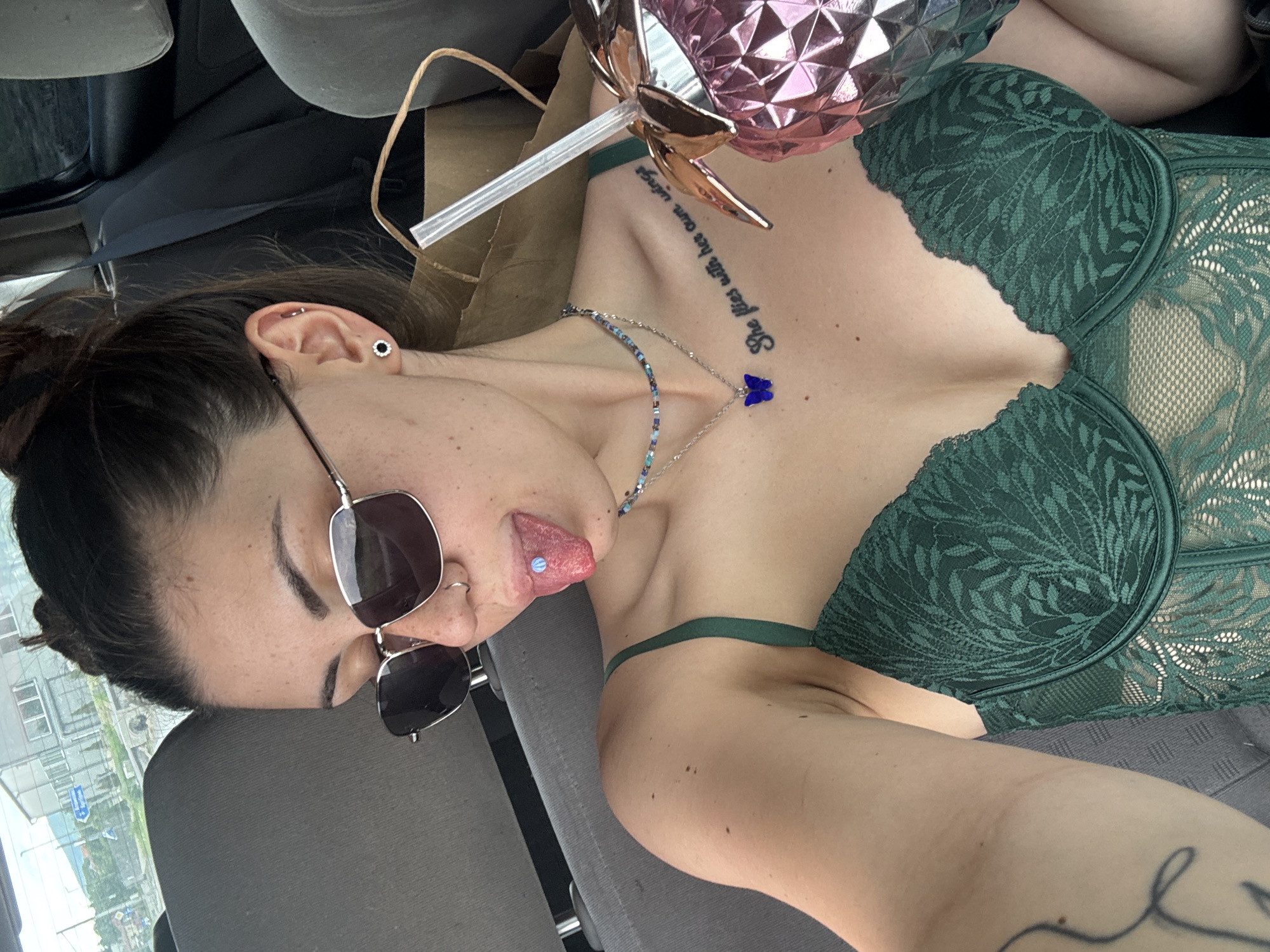 An adult female is pictured in a car, reclining in the passenger seat, oriented on her side with her head to the left of the frame. She is wearing a dark green lace bralette or bustier. Her dark hair is pulled back, and she is wearing sunglasses pushed up on her nose, a silver ring in her nose, and multiple ear piercings, including a black floral stud in her earlobe and a silver ring in her helix. A blue beaded necklace with a prominent blue butterfly charm hangs around her neck.
She is sticking her tongue out, revealing a light blue or white stud piercing. On her right collarbone, a tattoo reads "She flies with her own wings." A shiny, pink, pineapple-shaped cup with a straw is visible near her shoulder. To her left, a brown paper bag is present. The interior of the car, including a gray car seat and a black seatbelt, is visible. Through the car window in the background, a blurry outdoor scene with buildings and what appears to be a street or road is visible, suggesting a daytime setting in an urban or suburban area. The image context places the location in Gorna Malina, Bulgaria. The overall mood conveyed is playful and relaxed.
