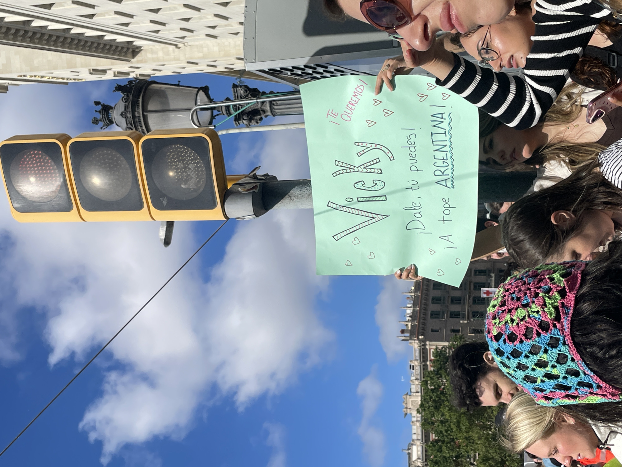 The image shows a group of people holding a sign in Barcelona, Spain. The sign is light green and has the name "Vicky" written in large letters. Above the name, it says "TE QUEREMOS!" followed by heart symbols, and below the name, it reads "Dale, tú puedes! ¡A tope ARGENTINA!". The people holding the sign appear to be supporters of the Argentine person named Vicky. In the background, there is a blue sky with clouds, a traffic light, a street lamp, and a building. The people in the foreground include some with knitted headbands, striped shirts, and sunglasses.