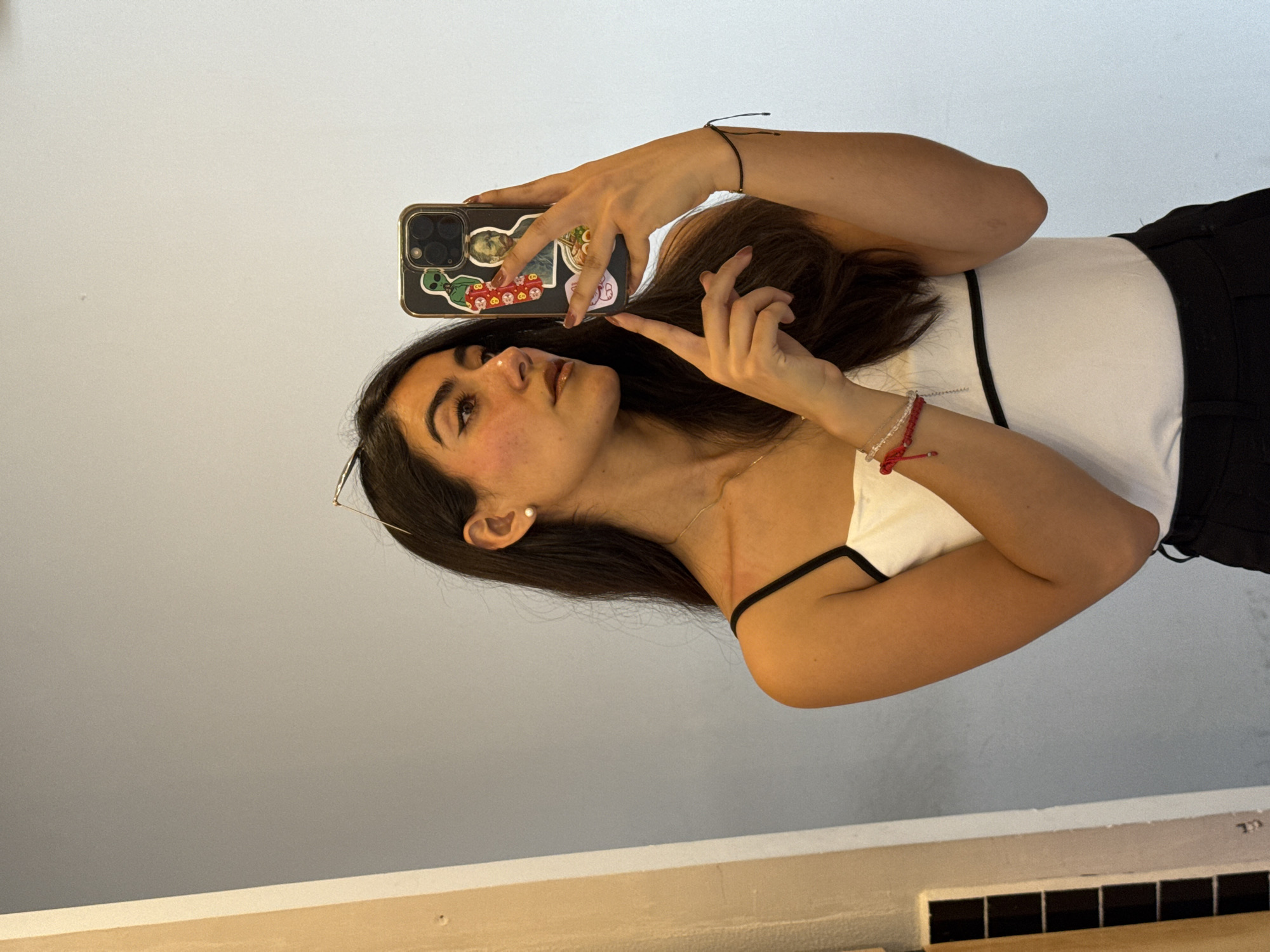 A young woman with dark hair and fair skin is taking a selfie with her phone. She is wearing a white tank top with black straps and black bottoms. The phone case has stickers on it. The location is likely indoors, with a beige wall visible in the foreground and a grey wall behind her. There is no indication of the location being in Puebla, Mexico.