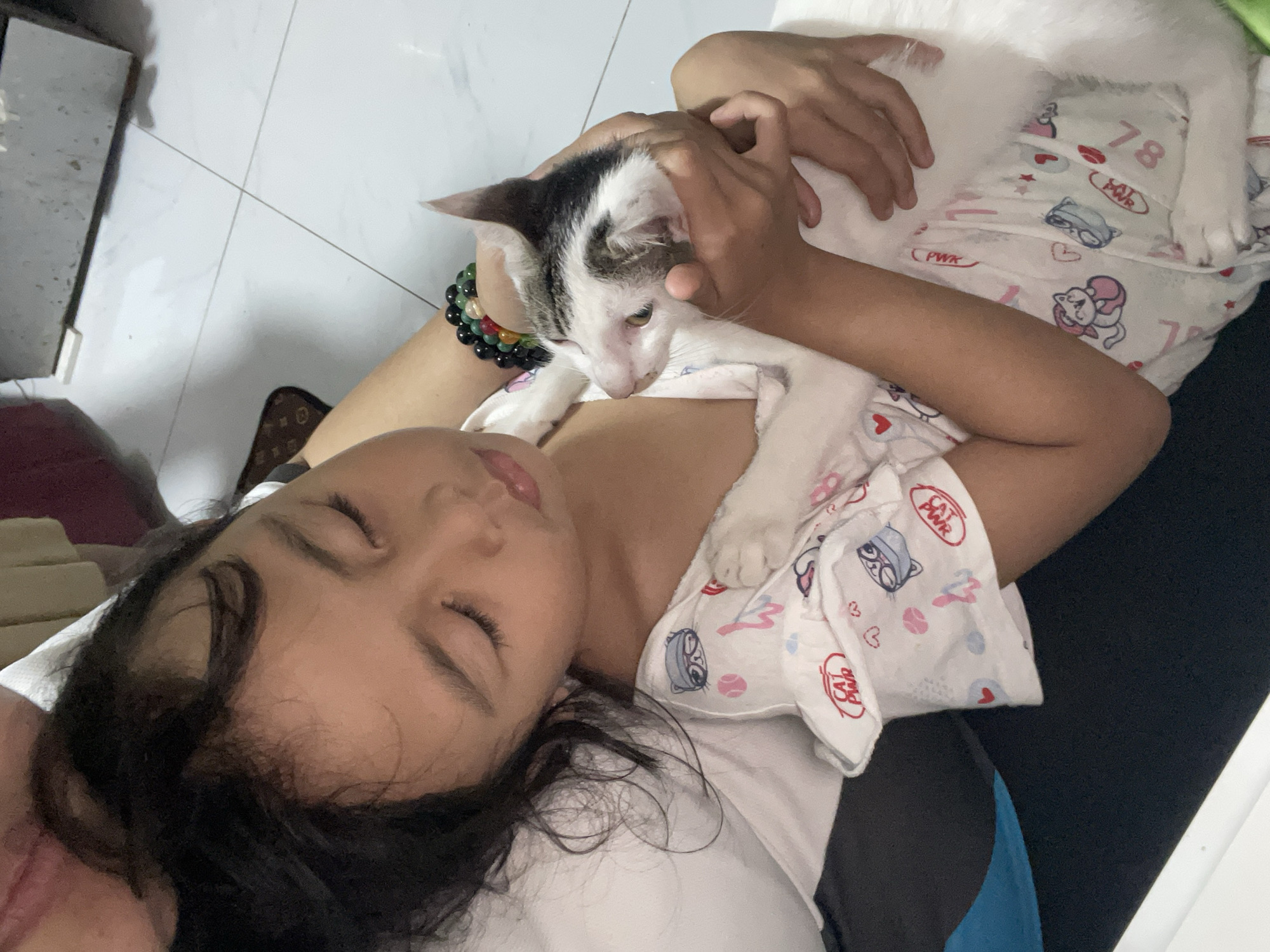 A young girl with dark hair and closed eyes is holding a white and black cat. She is wearing pajamas with cat-themed prints. The cat is being held securely in her arms. The background shows a tiled floor and a glimpse of furniture. The scene appears to be indoors, likely a home in Davao City, Philippines.