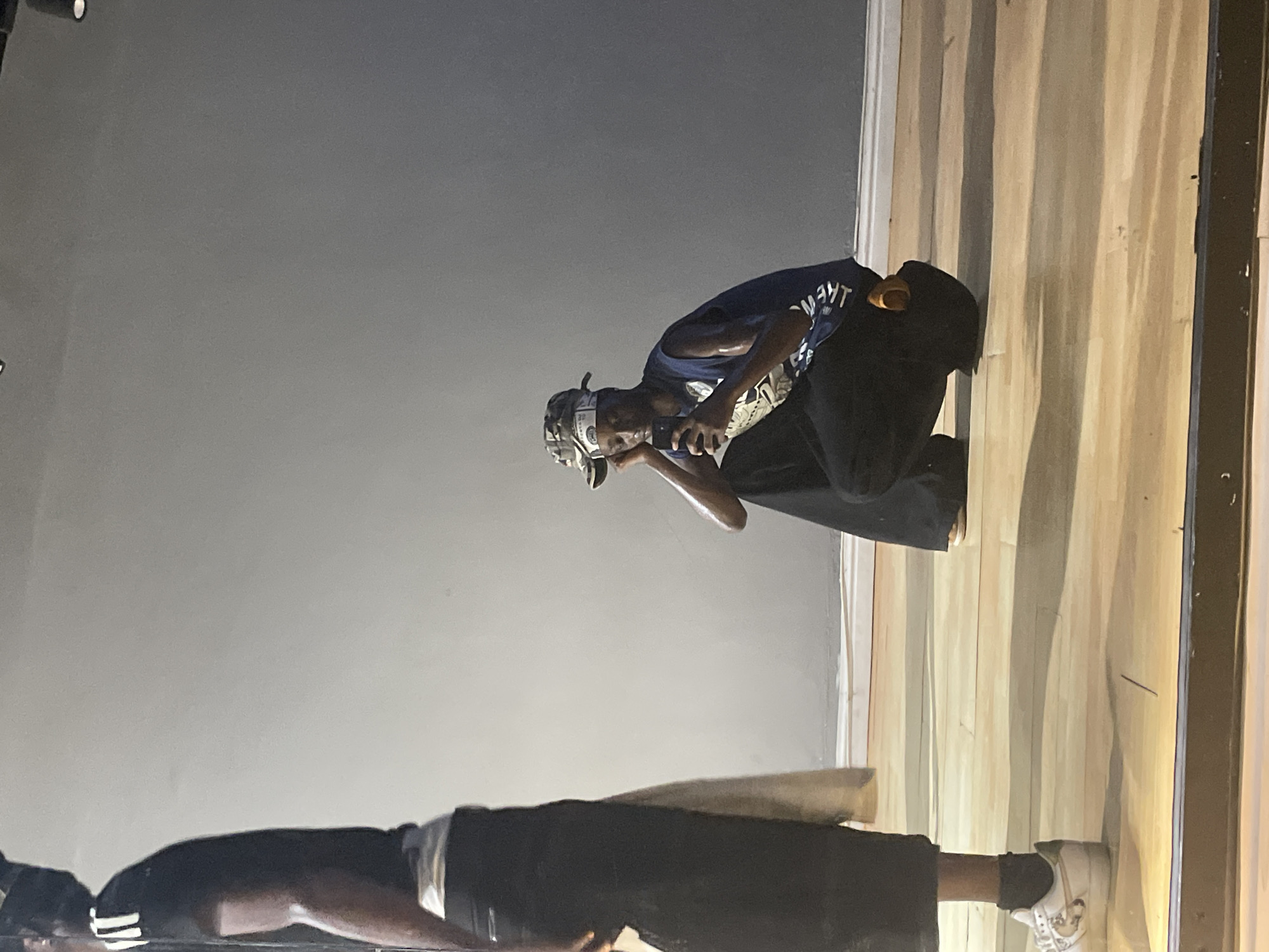 This image, taken in Lekki, Nigeria, features two individuals in an indoor setting, likely a studio or stage, with a perspective from slightly above.
In the middle ground, a person with dark skin is captured in a crouching or kneeling pose on a polished wooden floor, looking downwards. They wear a dark blue t-shirt with a white graphic or text that appears inverted and partially visible as "THEW...". A bandana or head covering adorned with dollar bill patterns is worn on their head. Dark pants complete their attire. The individual is holding a small, dark object, resembling a phone or camera, in their hands, pointing it forward and downwards.
The foreground prominently displays the lower body of another person, or a strong reflection, on the highly reflective wooden floor. This individual wears dark clothing, white socks, and white sneakers with dark details, possibly stripes. The glossy surface of the wooden planks clearly shows a distorted reflection of an upper body. The background is a plain, light gray wall, which provides a neutral backdrop to the scene. The artificial lighting creates some subtle shadows, and the overall atmosphere is focused and calm.