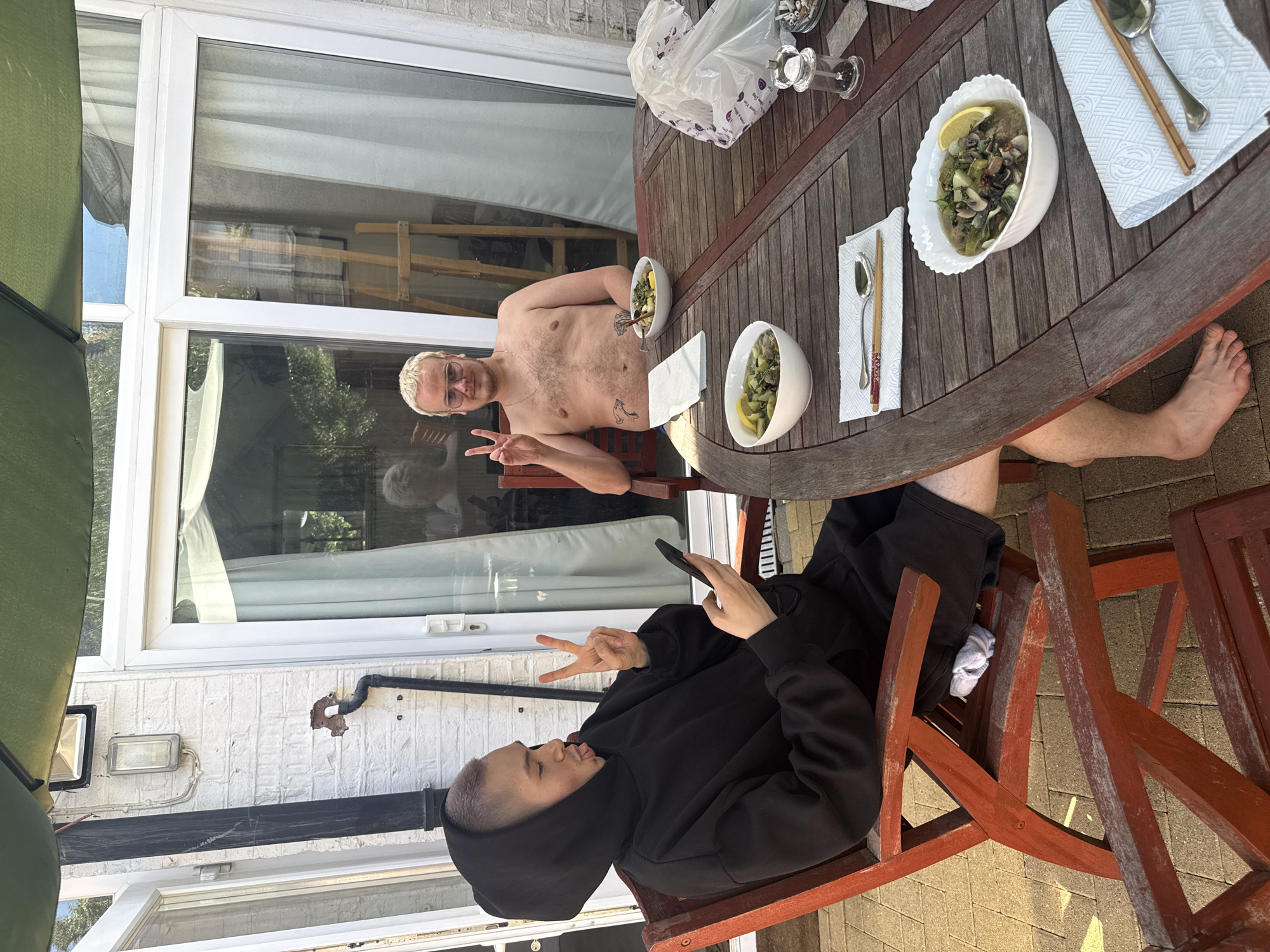 Two individuals are seated at a wooden table outdoors, situated on a patio with brick paving. The background features a white brick building with large glass doors. The overall impression is a casual meal in a residential setting.
The person on the left is wearing a black hooded sweatshirt and is looking at their phone while holding it in their left hand. Their right hand is raised, displaying a peace sign. They are sticking their tongue out slightly and have light-colored hair.
The person on the right is shirtless and wearing glasses. They are also making a peace sign with their right hand. Both individuals appear to be enjoying a meal.
On the table, there are two white bowls containing food, possibly a stew or soup with vegetables and mushrooms, each garnished with a lemon slice. Cutlery, including chopsticks and spoons, is arranged on napkins beside the bowls. A plastic bag and a pepper grinder are also visible on the table. The wooden chairs are red and appear to be weathered. The environment suggests a sunny day.