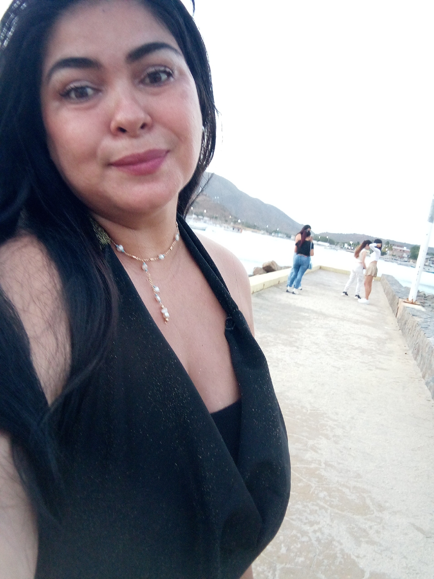 The image is a selfie of a woman with long black hair, wearing a black top and a delicate beaded necklace. In the background, several people stand on a concrete pathway near the water's edge. Behind them is a bay surrounded by low hills or mountains. The setting appears to be a harbor area in Juan Griego, Venezuela, based on the architecture and landscape.