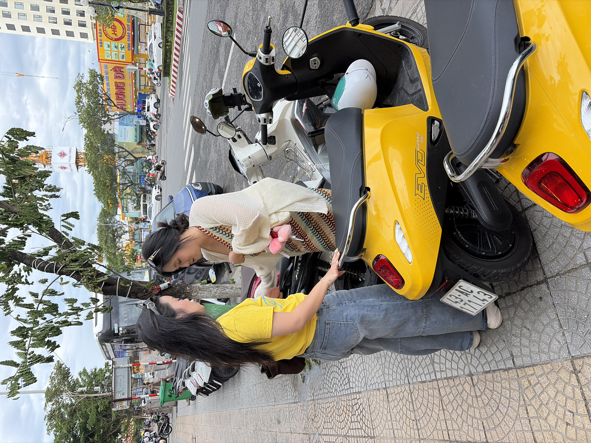 Two young Asian women are standing on a sidewalk in Da Nang, Vietnam, next to two parked scooters. The woman on the left, wearing a bright yellow t-shirt and light blue jeans, is facing the other woman and appears to be talking to her. She has long black hair and is wearing sunglasses on her head. The woman on the right, dressed in a white cardigan over a patterned dress, is also facing the first woman, looking down. She has her hair pulled back and is also wearing sunglasses on her head.
The scooters are parked along the sidewalk, with a yellow scooter prominently in the foreground. The yellow scooter has a license plate that reads "43-AC 171.43". Behind it is a white scooter. In the background, traffic is visible on a street, including cars and other scooters. Buildings line the street, some with awnings and signage. Trees with green foliage are present along the sidewalk and in the background, and a cloudy sky is overhead. A green trash bin is also visible near the scooters. The sidewalk is paved with interlocking patterned tiles.