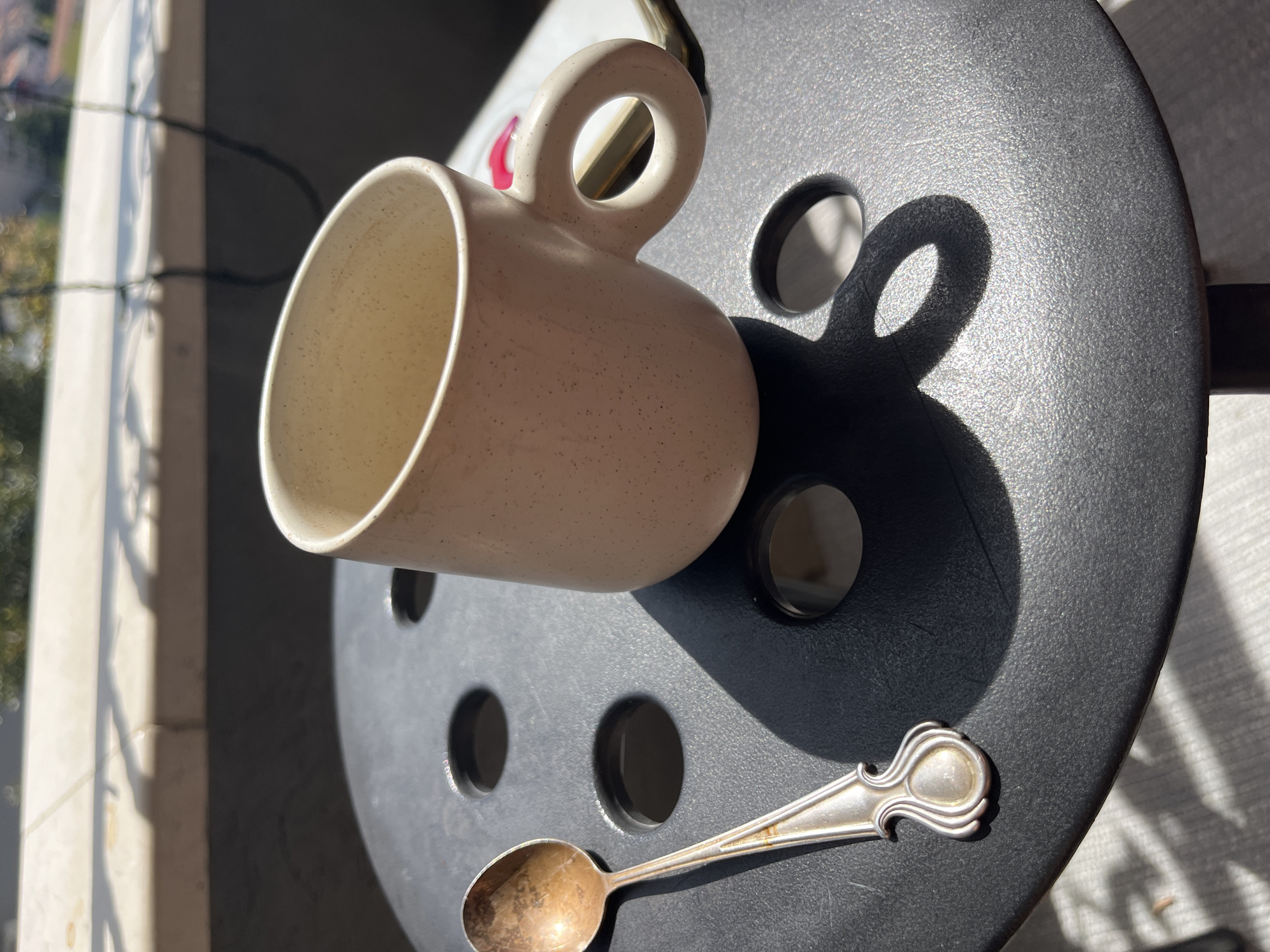 A beige ceramic mug with a round handle sits on a dark gray, perforated, round table. A tarnished silver spoon lies next to the mug. The scene is illuminated by sunlight, casting shadows of the mug and table features. In the background, a balcony and exterior elements are visible, suggesting an outdoor setting. The location is consistent with a cafe or outdoor space in a sunny locale. There's no evidence to suggest the image was taken in Budva, Montenegro. The location cannot be definitively ascertained from the given information.