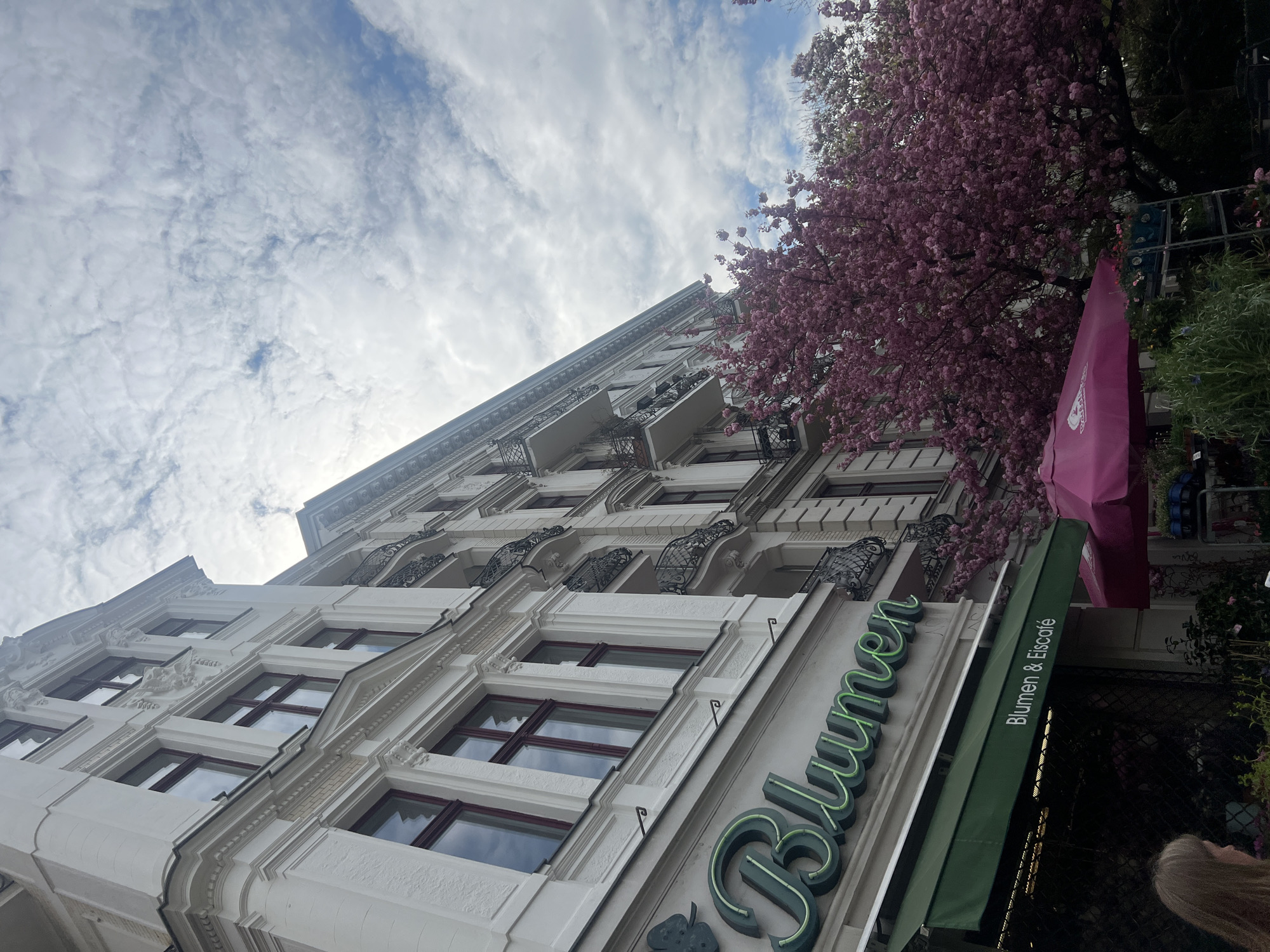 The image shows the exterior of a building in Berlin, Germany, specifically the "Blumen & Eiscafe" (Flowers & Ice Cream Cafe). The building is several stories high, with white walls, dark-framed windows, and decorative wrought-iron balconies. A green awning above the entrance displays the cafe's name. A tree with pink blossoms is visible on the right side of the building. The sky above is cloudy.