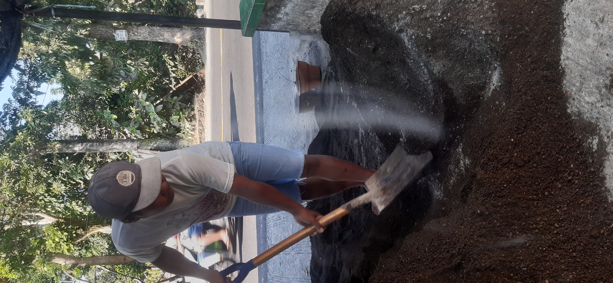 A person, wearing a white t-shirt, denim shorts, and a dark baseball cap, is using a shovel with a blue handle to mix a pile of dark soil. The person's face is obscured by the cap. The action is taking place outdoors, with lush green trees visible in the background and a paved road with a yellow line running through it. Two small pots, one brown and one dark gray, are situated behind the pile of soil, near a low concrete structure with a green trim. The lighting suggests it is daytime, with sunlight casting shadows. The context implies an activity related to gardening or construction. The location is identified as Sepakek, Indonesia.