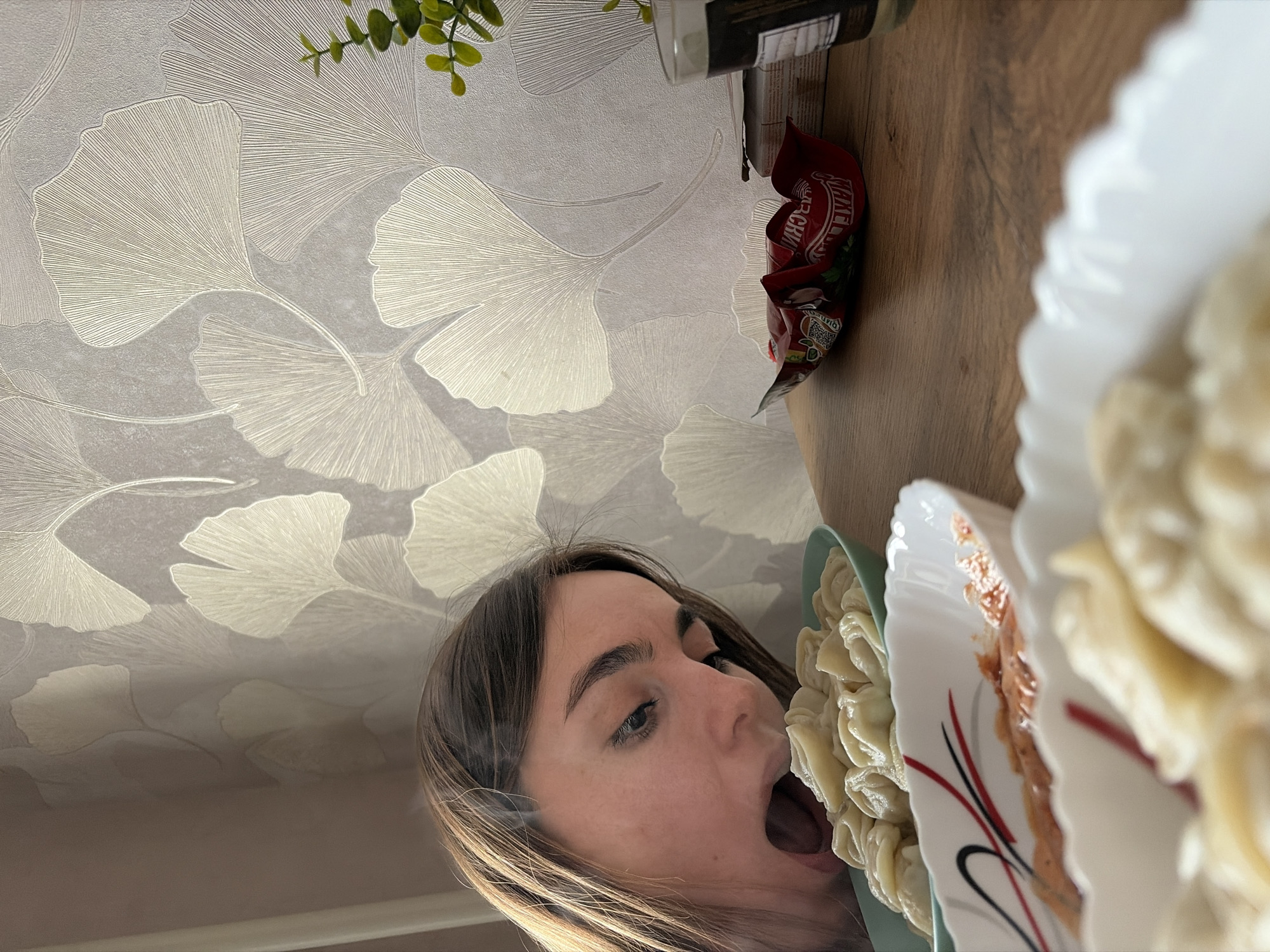 The image shows a woman with brown hair and eyes, mouth open, appearing to eat dumplings. The scene is indoors, with the ceiling featuring a wallpaper pattern of ginkgo leaves. On a wooden surface, there is a bag of "Mikoyan" dumplings, a can or bottle obscured from clear view, and plates of dumplings, one with sauce. There is no clear indication of the location in the city of Izhevsk, Russia.