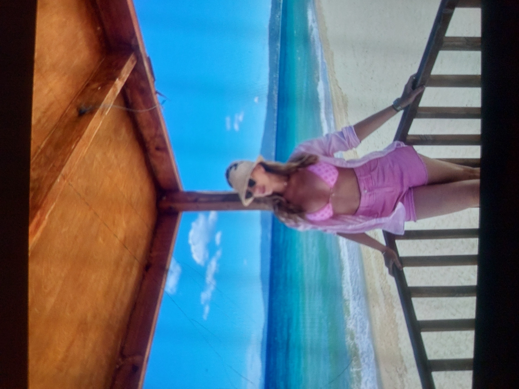 A woman in a pink bikini and shorts with a hat and sunglasses stands on a wooden ladder next to a beach. The beach is on the coast of Tsarevo, Bulgaria. The woman is holding onto the ladder with one hand. The scene is viewed from inside a wooden structure. The sky is blue with some clouds. The water is turquoise. The beach is white sand.