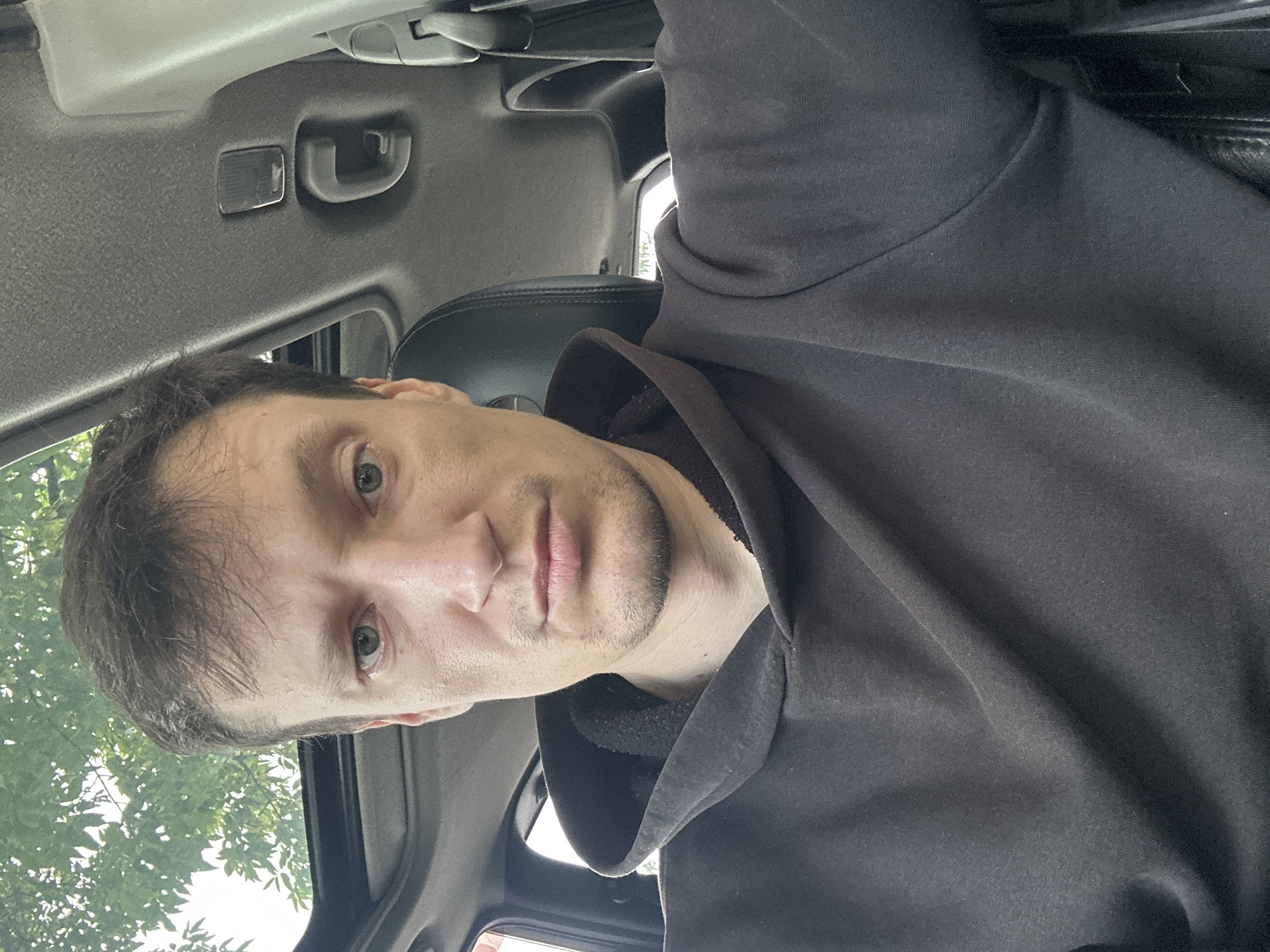 A medium shot selfie shows a fair-skinned man in his late 20s or early 30s. He has short brown hair, light blue eyes, and a clean-shaven face with a light stubble around his chin. He is wearing a dark brown hooded sweatshirt. The man is sitting in the passenger seat of a car, with the sunroof open. The interior of the car, including the headliner and a portion of the driver's seat, is visible. Trees and foliage are seen through the sunroof, suggesting an outdoor location. The photo appears to have been taken during the daytime. The location is identified as Makiv, Ukraine.