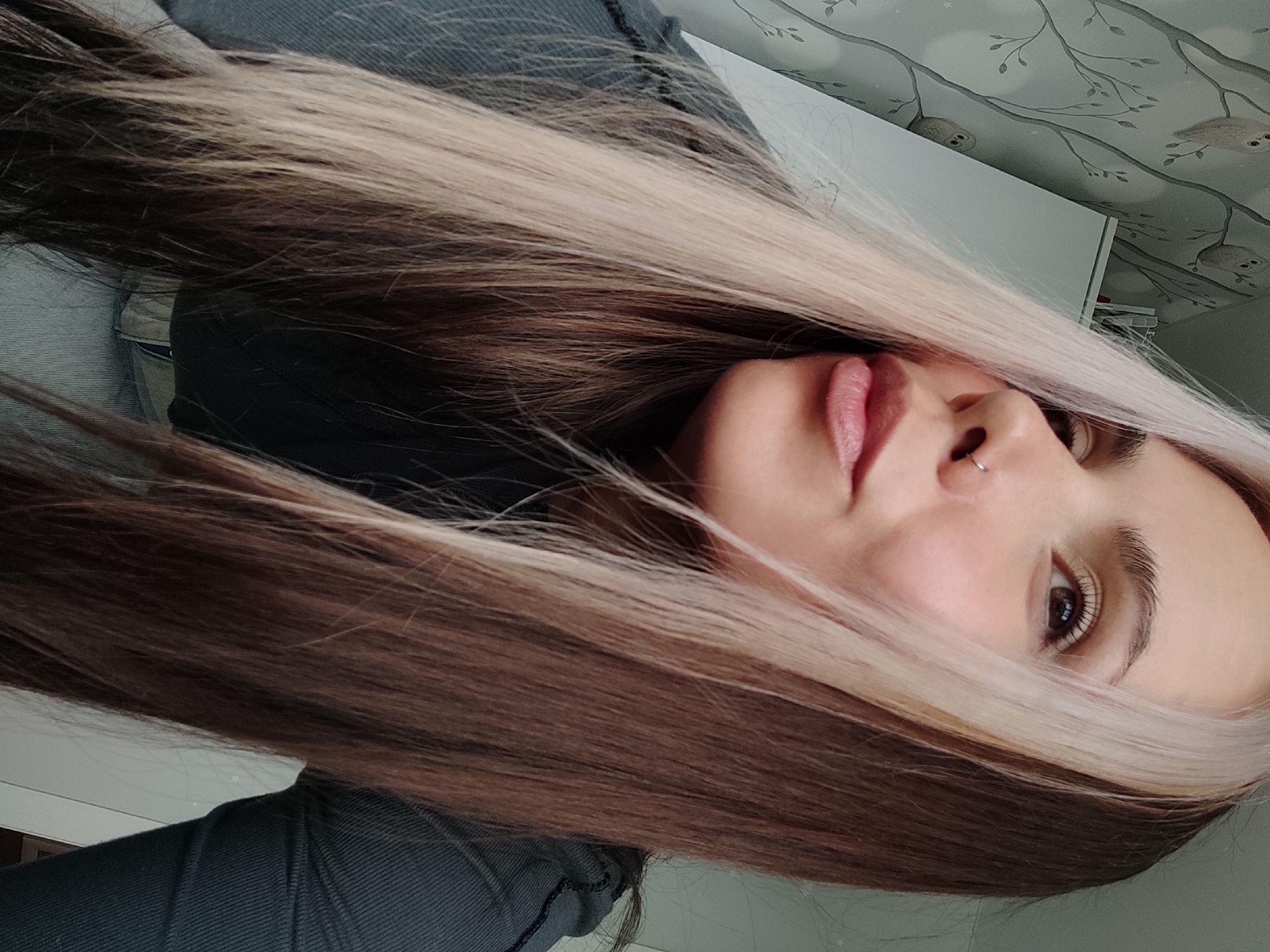 This close-up portrait-style image, taken from a rotated perspective, features a young person with long, flowing hair. The subject's face is partially obscured by their hair, with the right eye, nose, and mouth in clear view. The hair displays a distinct two-tone coloring: dark brown or auburn on the underside, transitioning to a bright blonde or platinum on the top layers, framing the face. A small, silver hoop nose ring is visible on the right nostril, and the lips are slightly parted. The person's right eye has visible eyelashes and a neutral, direct gaze. They are wearing dark, possibly gray, textured clothing, resembling a sweater or hoodie.
The background indicates an indoor setting. To the upper right of the person's head (as oriented in the image), a section of light-colored furniture, possibly a desk or shelf, is visible. Above this, the wall is covered with a light-toned wallpaper featuring a pattern of tree branches and cartoonish owls. No specific activity is depicted beyond posing for the photograph. There is no visible text. The setting is in Chaikovsky, Russia.