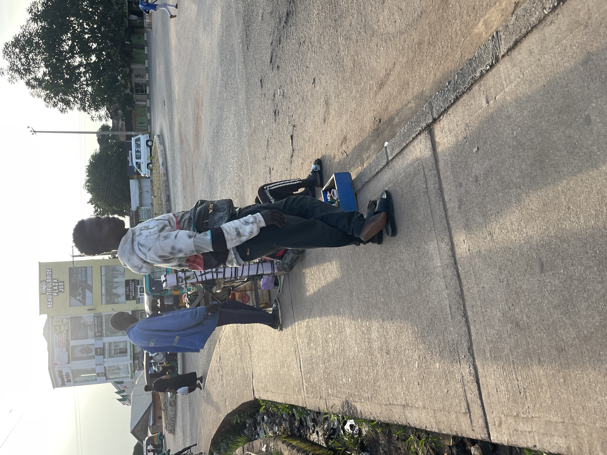 The image depicts a street scene in Jos, Nigeria, near the "Beautiful Bride Plaza." In the foreground, a young Black man, wearing a grey hoodie with white and red accents and black pants, stands on a concrete sidewalk, facing away from the camera and looking towards the right. He has a black crossbody bag slung over his shoulder. He is wearing black slides. To his left, a person in a blue jacket and dark pants is also standing on the sidewalk, facing away from the camera. Further to the left, a woman in a dark dress walks away from the camera, carrying a white bag. A white van is parked in the background. The street appears to be a commercial area with several buildings, including the aforementioned plaza, which has signage indicating it offers services related to weddings and jewelry. The environment appears somewhat dusty, with a mix of paved and unpaved surfaces. The overall lighting suggests it is either early morning or late afternoon.