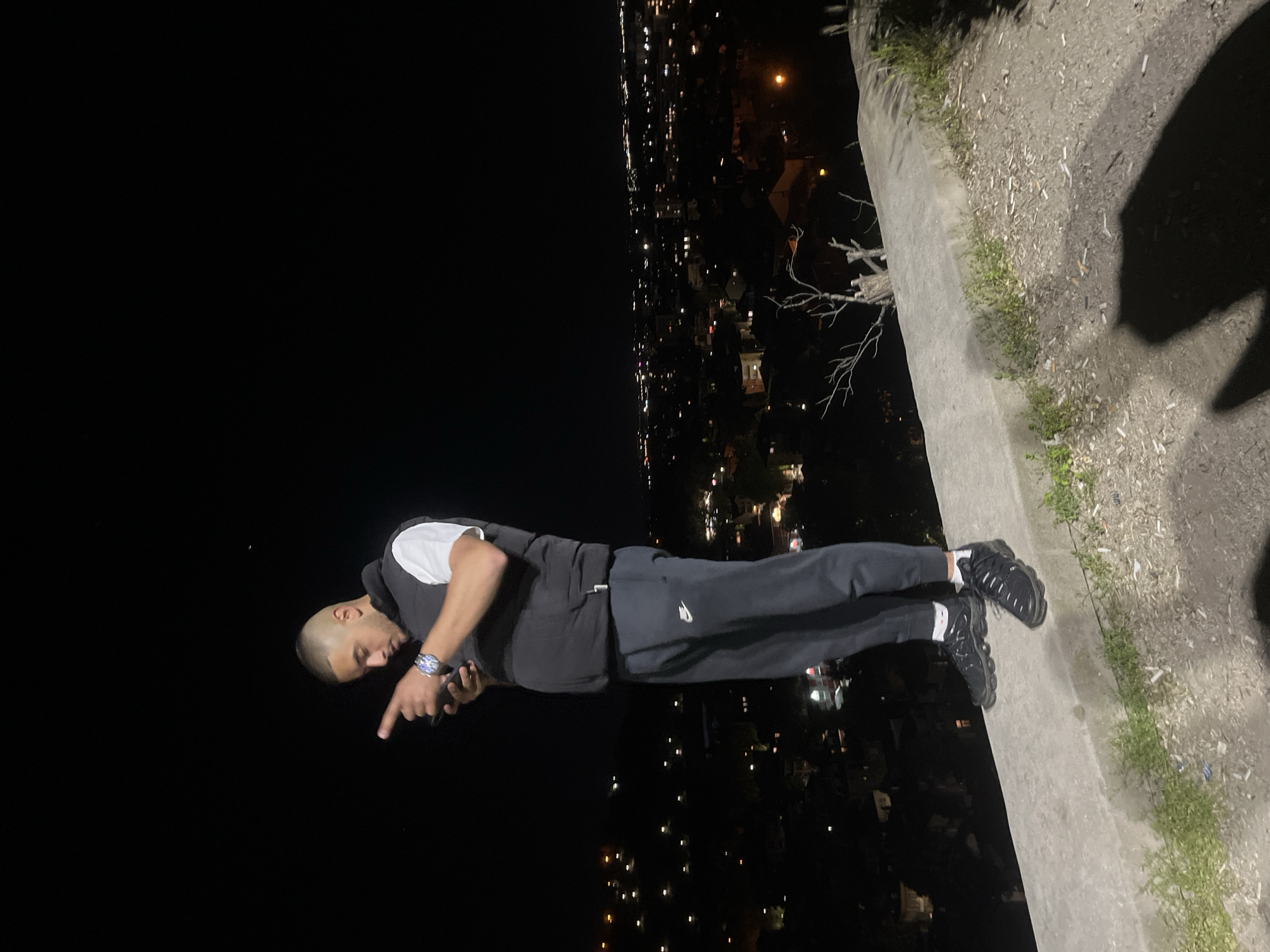 A person is standing on a ledge overlooking a city at night. The person has a shaved head and wears a white t-shirt with a black vest and dark trousers. They are pointing downwards with their right hand, holding a dark-colored object (likely a phone) in their left hand. The city below is illuminated with scattered lights. The ledge appears to be made of concrete and has some vegetation growing along it. It is not possible to verify the location as Plovdiv, Bulgaria, based on the image alone.