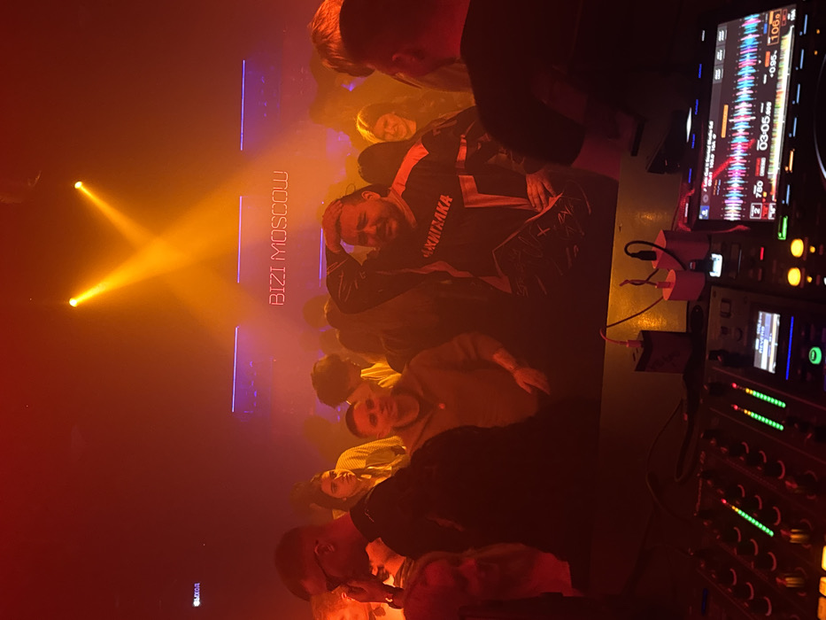 The image portrays an interior scene within a nightclub or music venue identified by a prominent neon sign displaying "BIZI MOSCOW" on the back wall, establishing the location as Moscow, Russia. An "ВЫХОД" (Exit) sign, partially visible on the far left wall, further confirms the Russian context. The environment is dark, filled with atmospheric smoke or haze, and illuminated by dominant red and orange lighting originating from two converging overhead spotlights and linear purple neon strips along the walls.
In the foreground, a DJ mixing console, identified as a Pioneer unit, occupies the lower portion of the frame. It features multiple illuminated faders, buttons, and a digital screen displaying audio waveforms, track timing (e.g., 03:05:00), and other music information. Two USB wall chargers, one black and one white, with cables attached, rest on the surface behind the DJ equipment.
The midground and background are densely populated by an estimated 15-20 individuals, primarily adults. A central male figure, possessing a beard and mustache, wears a black and orange long-sleeved shirt with the Cyrillic text "АКТИВАКА" (Aktivaika) printed on it. This individual holds his right hand to his forehead, looking forward with a discernible expression. To his left, another male in a dark hoodie faces forward. Various other individuals, both male and female, are visible throughout the crowd, standing, observing the scene, or engaged in social interaction. One female individual in the background exhibits a smile. The overall activity suggests a social gathering at a musical event.