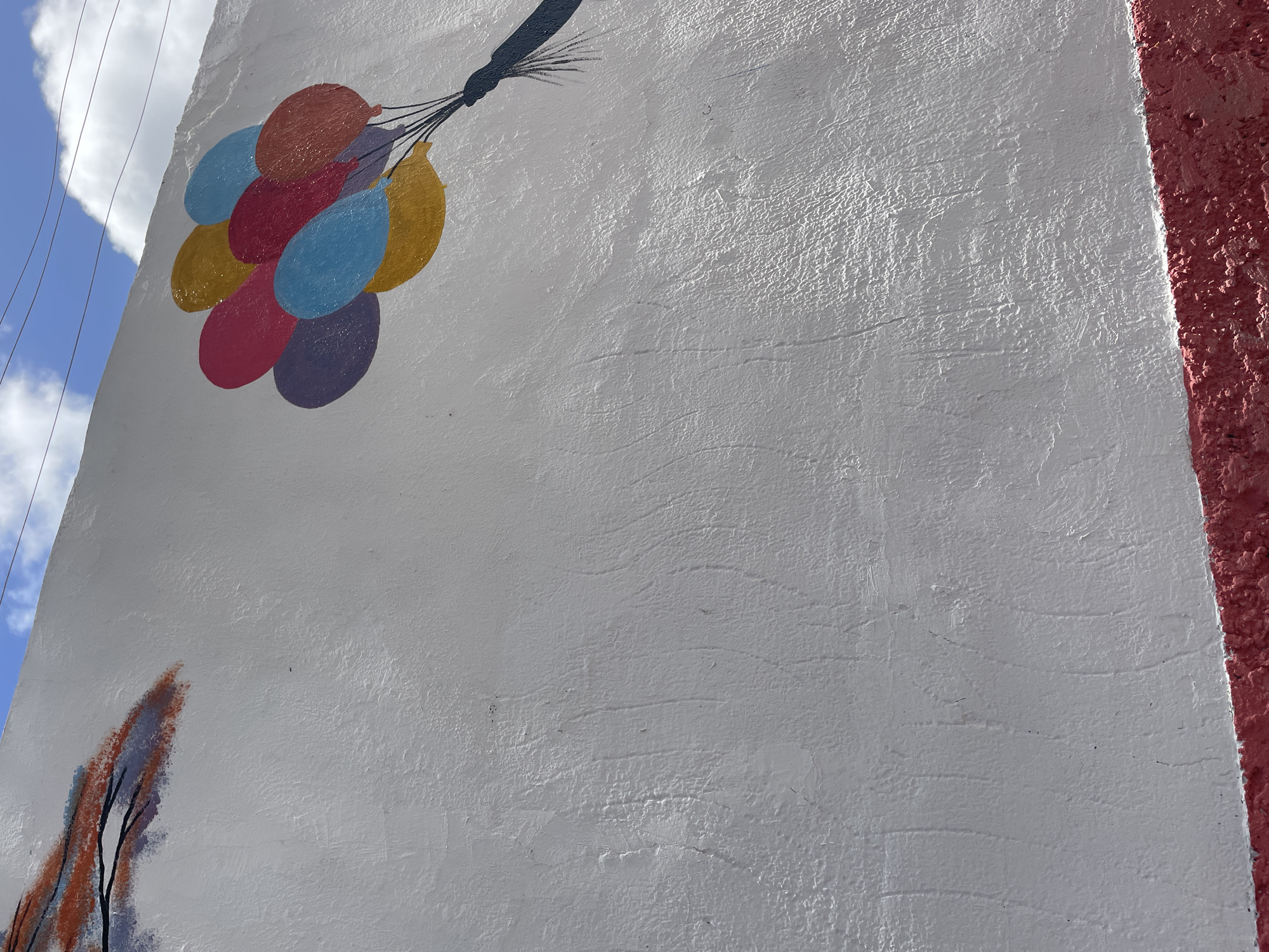 The image shows a white stucco wall with a mural. In the upper left, a depiction of a tree branch with reddish-orange and blue-gray leaves is visible. To the right, a cluster of colorful balloons – purple, red, yellow, and blue – are painted, appearing to float upwards, tethered by black strings to a dark blue string holder that extends out of the frame. The wall is textured, and the blue sky with white clouds is visible at the top. A red, textured border runs along the bottom of the wall. This mural is located on a wall in El Valle del Espíritu Santo, Venezuela.