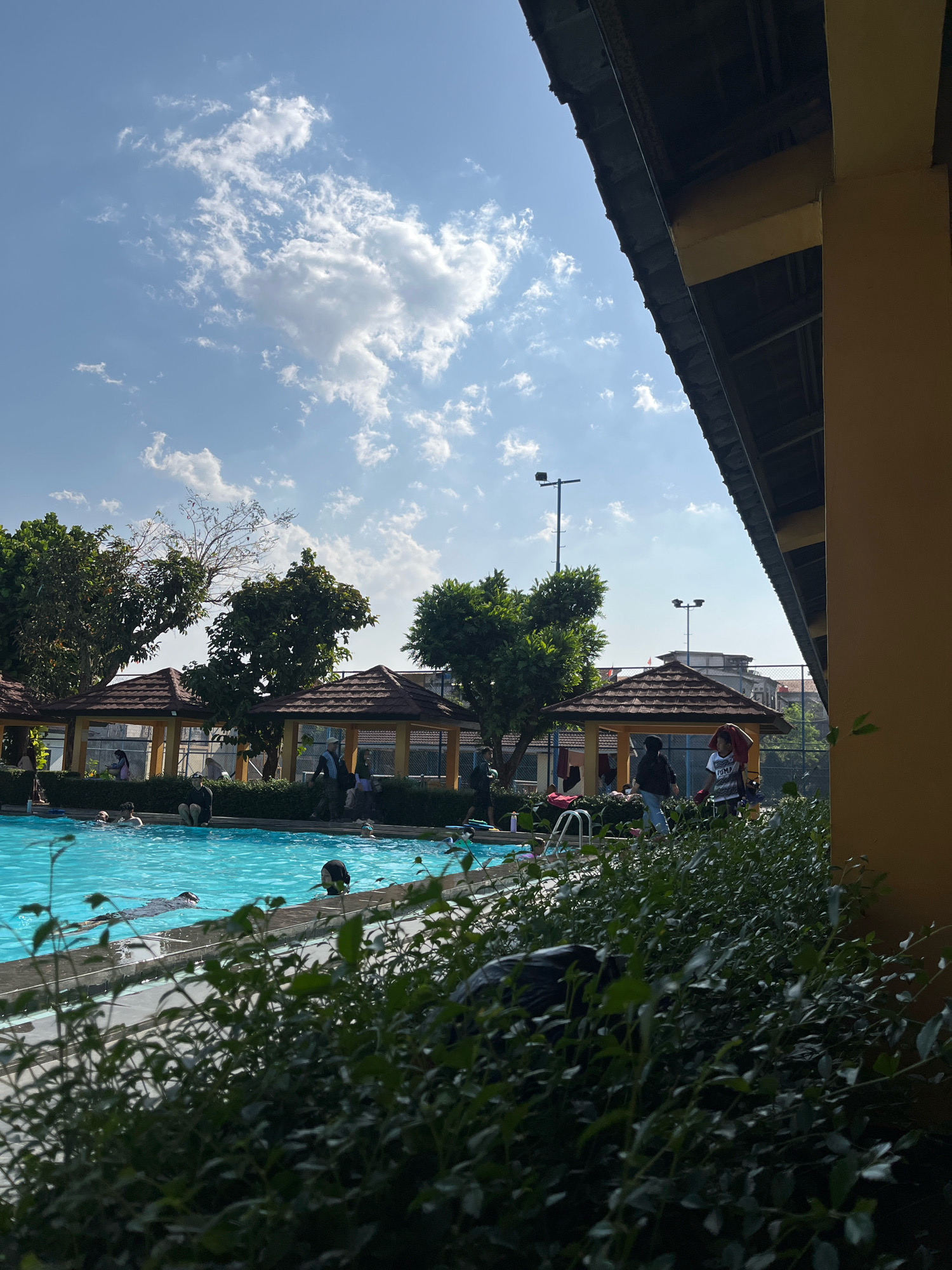 The image depicts an outdoor swimming pool area in Sariwangi, Indonesia, under a bright, partially cloudy sky. The foreground is dominated by lush green foliage, partially obscuring the view of the pool and its surroundings.
In the mid-ground, a clear blue swimming pool is visible, with several individuals swimming and wading. Along the edge of the pool, a gray tiled border is present, and a set of silver pool ladders are installed. Beyond the pool, a row of open-sided gazebos with brown tiled roofs provides shade. Several people are seen gathered or walking around these gazebos and the poolside. Tall green trees with dense foliage are scattered throughout the scene.
In the background, a tennis court fence can be observed, and beyond that, buildings with red-tiled roofs are visible. Two tall lampposts are also present in the background, suggesting the area is equipped for evening use. The right side of the image features a portion of a building with a bright yellow exterior and a dark overhanging roof structure. The overall impression is a recreational area on a sunny day.