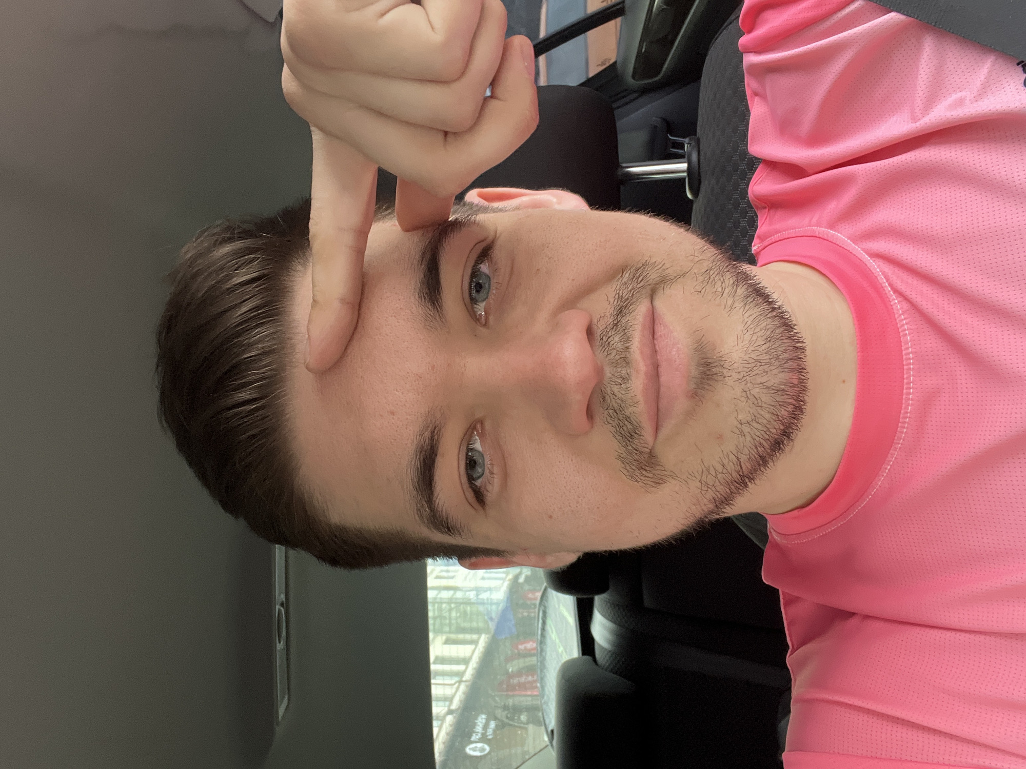A close-up, rotated selfie of a Caucasian male with brown hair and blue eyes, wearing a pink athletic shirt, likely inside a vehicle. His right index finger is touching his forehead. Outside the vehicle window, partial visibility of a street in Yekaterinburg, Russia, is observed, evidenced by Russian signage. Interior is dark.
