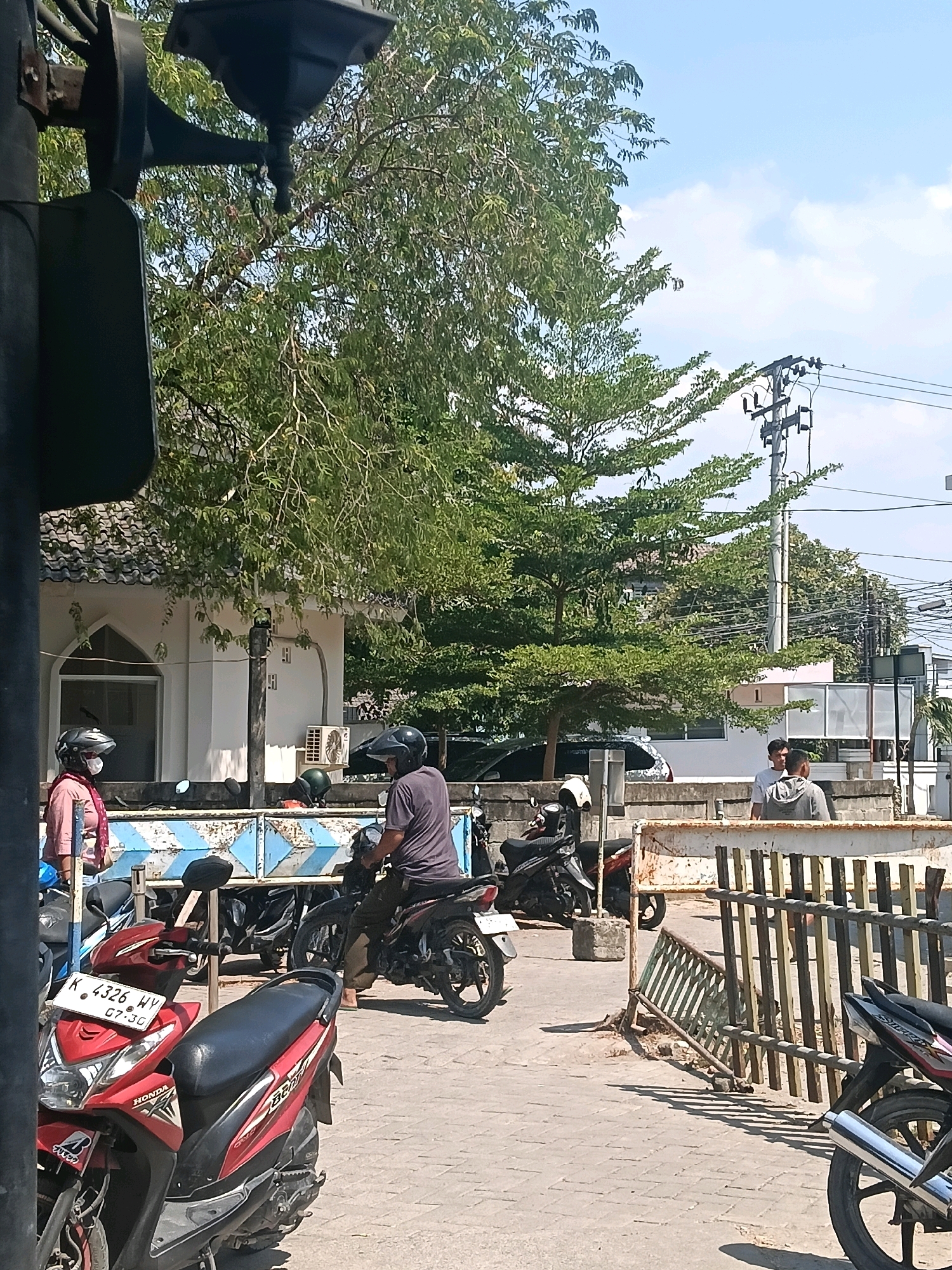 This image depicts an outdoor scene in Balun, Indonesia, on a sunny day. In the foreground, a red Honda motorcycle is parked, with its license plate reading "K 4326 HY". Behind it, another motorcycle, partially obscured, is visible. Two individuals wearing helmets are present; one, in a pink shirt and scarf, stands near the red motorcycle, while another, in a dark grey shirt, sits on a motorcycle in the mid-ground. A white building with a tiled roof, possibly a mosque or community center, is situated in the background, with an air conditioning unit mounted on its exterior wall. Several other motorcycles are parked in the vicinity. A metal barrier, made of vertical bars and a horizontal railing, separates the area. Trees with lush green foliage dominate the upper portion of the image, with power lines and utility poles visible against a blue sky with scattered clouds. The ground is paved with interlocking bricks.