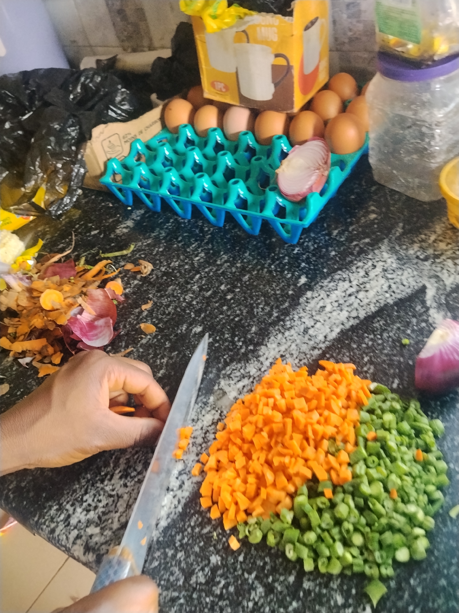 This is a close-up, overhead shot of a person's hands preparing vegetables on a speckled granite countertop. The scene appears to be in a kitchen. In the foreground, a hand is holding a knife, in the process of chopping carrots. A pile of chopped carrots and green beans is visible on the counter. To the left, there is a pile of vegetable scraps, including carrot peels and onion skins.
In the background, a blue egg carton holds several brown eggs. A sliced red onion is also placed within the egg carton. Behind the eggs, a yellow cardboard box with an image of a mug is partially visible. To the right of the eggs, a large, clear plastic container with a purple lid is filled with what appears to be cooking oil or some other liquid. A black plastic bag filled with trash is situated to the left of the egg carton.
The lighting suggests it might be daytime, but no specific time can be determined. There are no obvious cues about the weather or external location. The overall impression is one of domestic food preparation. There is no visible text in the image.