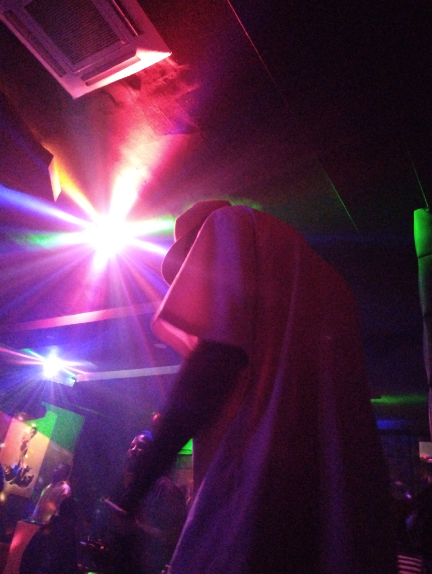 The scene appears to be inside a nightclub or entertainment venue, possibly located in Gombe, Nigeria. The lighting is colorful and intense, featuring bright spotlights with green, blue, purple, and red hues. The person in the foreground wears a light-colored top. Multiple individuals stand in the background, some holding objects. The environment includes a ceiling-mounted air conditioning unit and a decorative green pillar.