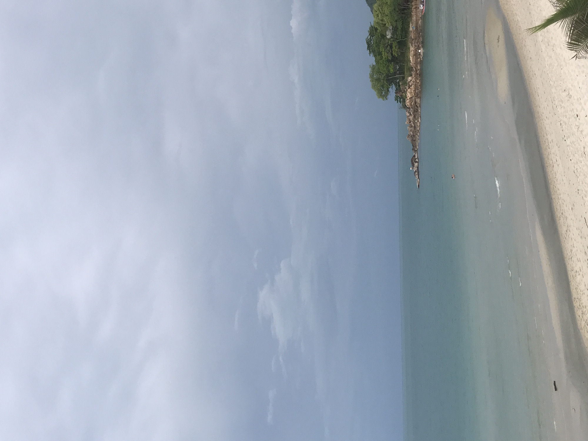 The image shows a beach scene, likely in Baan Chaweng, Thailand. The sky is light blue with scattered clouds. The ocean is a pale turquoise color, meeting the sandy beach. A rocky shoreline with green vegetation and trees is visible along the edge of the water. A few small figures appear to be in the water. A palm tree is visible to the right of the frame.
