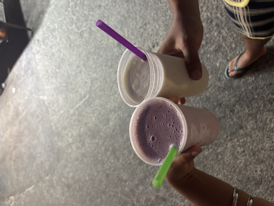 Two people hold up cups of drinks with straws in them. One cup is a light yellow color, and the other is a light purple color. The straws are purple and green, respectively. The drinks are held in the hands of two different people. One is wearing black and white plaid shorts and blue flip flops. There is a concrete floor in the background.