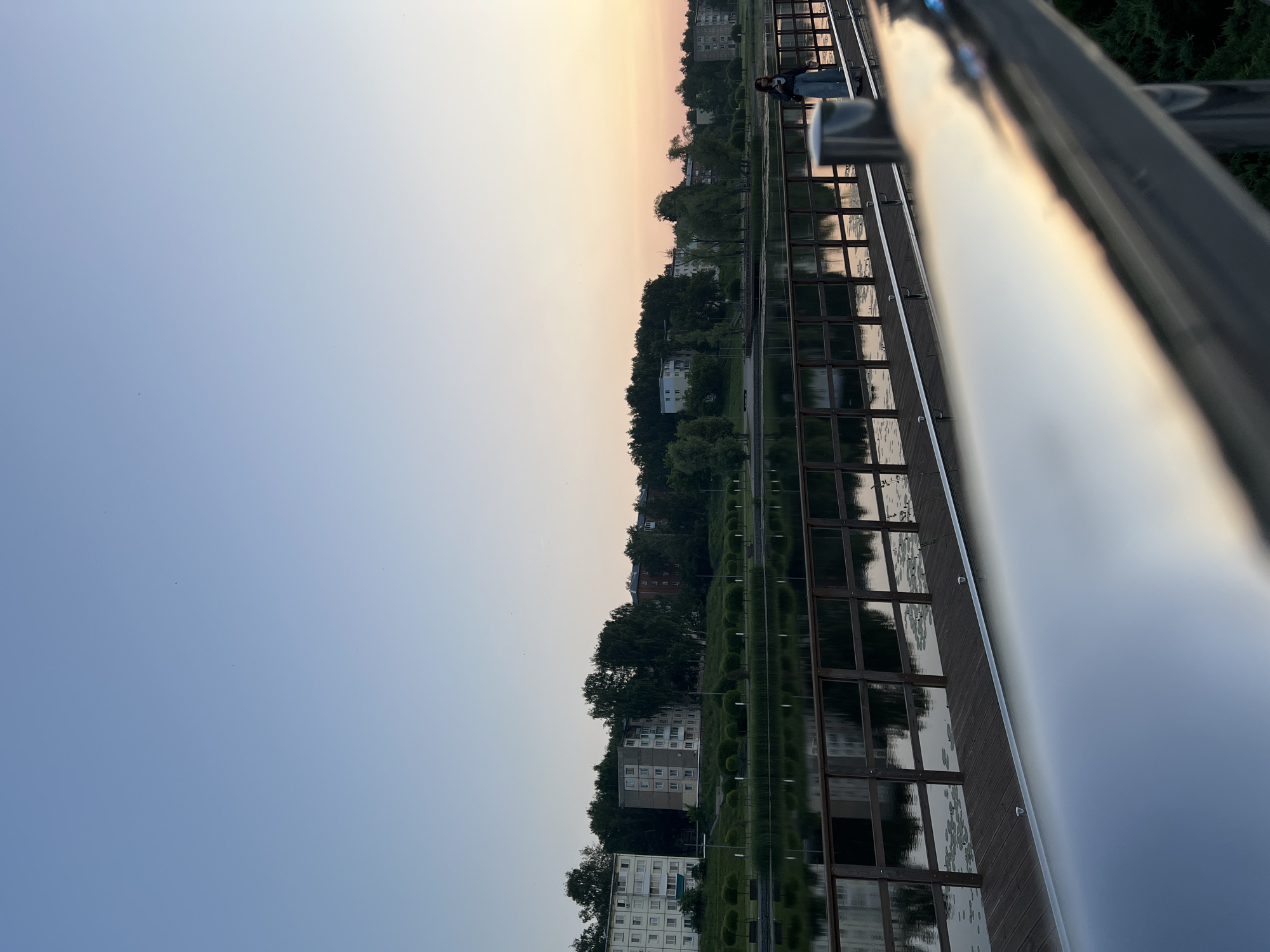 This is a horizontal, eye-level, medium shot of a wooden walkway with a metallic railing in the foreground. The walkway extends across the lower half of the image and overlooks a calm body of water that reflects the buildings and trees lining the opposite bank. The sky above is clear and transitions from a light blue at the top to a soft orange and pink hue near the horizon, indicating either sunrise or sunset.
On the far bank, a line of trees with lush green foliage separates a row of apartment buildings from the water. The buildings are of varying heights and architectural styles, common in a Russian urban setting. The water surface is mostly still, with some lily pads visible.
On the walkway, a young woman with dark hair is standing near the railing on the right side of the image. She is wearing a dark jacket and light-colored pants and is looking down, holding what appears to be a phone. Her posture suggests she is either admiring the view or engaged with her device.
The metallic railing in the foreground is highly reflective, mirroring the sunset colors and creating a blurred, abstract effect that emphasizes the sunset. The overall atmosphere of the image is peaceful and serene, capturing a moment of quiet contemplation by the water. This scene is likely located in the city of Votkinsk, Russia, given the characteristic apartment buildings and the presence of a walkway and body of water common in such cities.