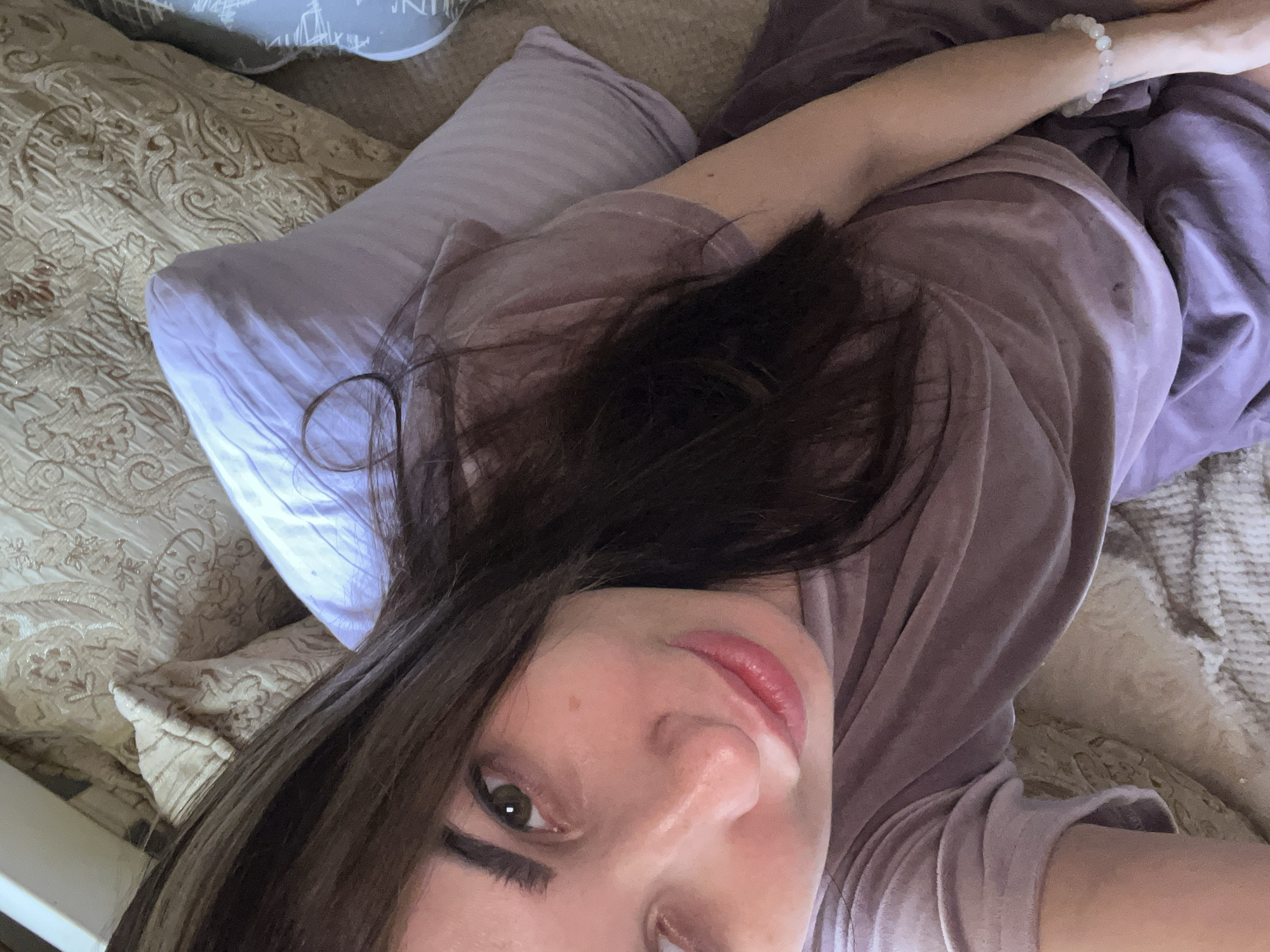 A close-up, overhead selfie shows a young woman with long, dark brown hair lying on a bed. She is wearing a light purple top and has a white beaded bracelet on her right wrist. Her eyes are green, and she is looking towards the camera with a slight smile. The bed appears to be covered in patterned pillows and blankets. The lighting is soft, and the overall impression is one of relaxation.