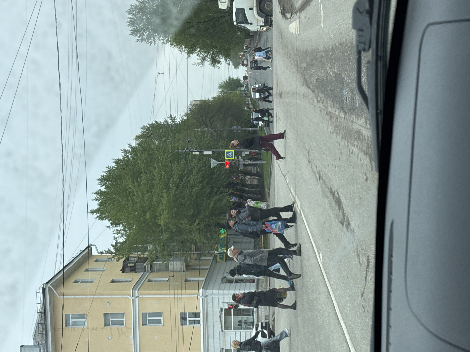 The image shows a street scene, likely taken from inside a vehicle in Perm, Russia. A group of pedestrians is crossing the street at a crosswalk. A traffic light is visible, and a yellow building is on the left side of the street. There are power lines and trees in the background. A truck is visible further down the road. The scene appears overcast.