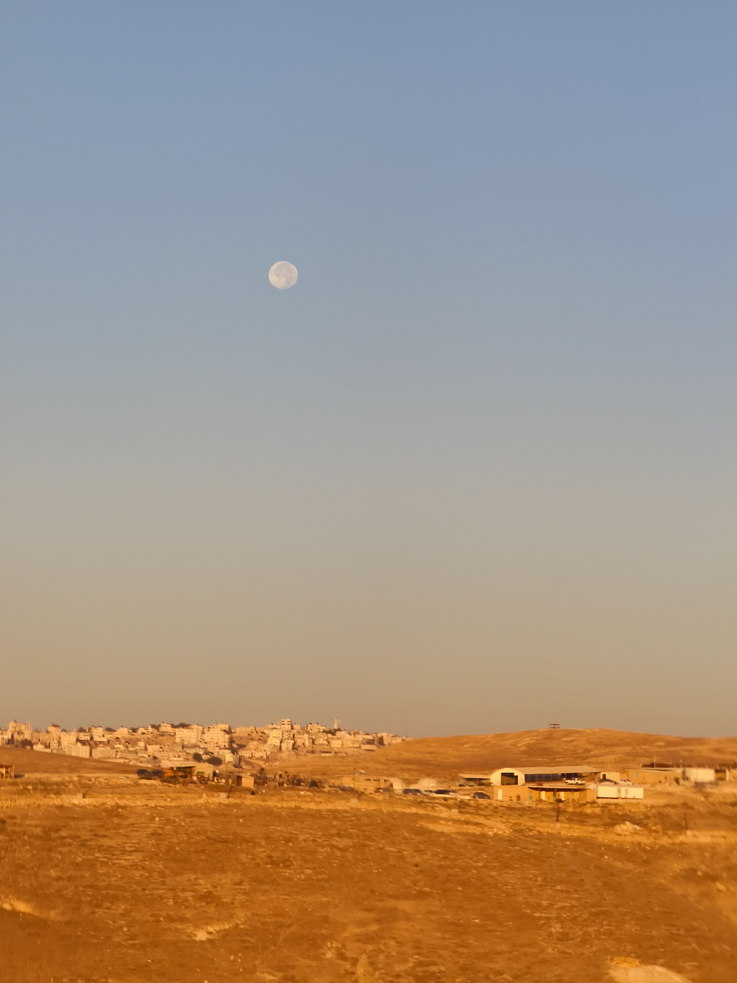 An eye-level, full outdoor shot captures a vast, undulating landscape bathed in the warm, golden light of sunset. In the distance, a town or village with light-colored buildings is clustered on a hill. The foreground is dominated by dry, golden-brown vegetation, with hints of green and darker brown, suggesting arid terrain. Scattered across the landscape are various structures, including what appear to be low-rise buildings with white roofs, some sheds, and a few vehicles. A prominent hill rises in the mid-ground, topped with a small, dark structure. High in the clear, pale blue sky, the full moon is visible, a luminous disc against the fading light. The scene conveys a sense of open space and a rural or semi-rural setting in the Palestinian Territories.