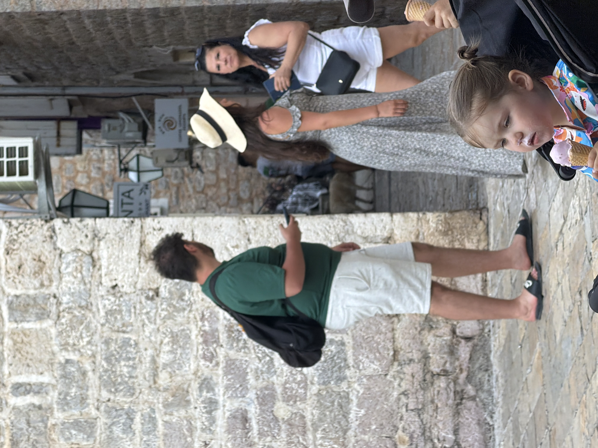 In Budva, Montenegro, a toddler with ice cream smeared on her face sits in a stroller. In the background, a man in a green shirt and white shorts carries a black backpack, holding a phone in his hand. To his right, a woman in a floral dress and a straw hat talks to another woman in a white top and white shorts. The scene is set against a backdrop of ancient stone buildings and cobblestone streets.