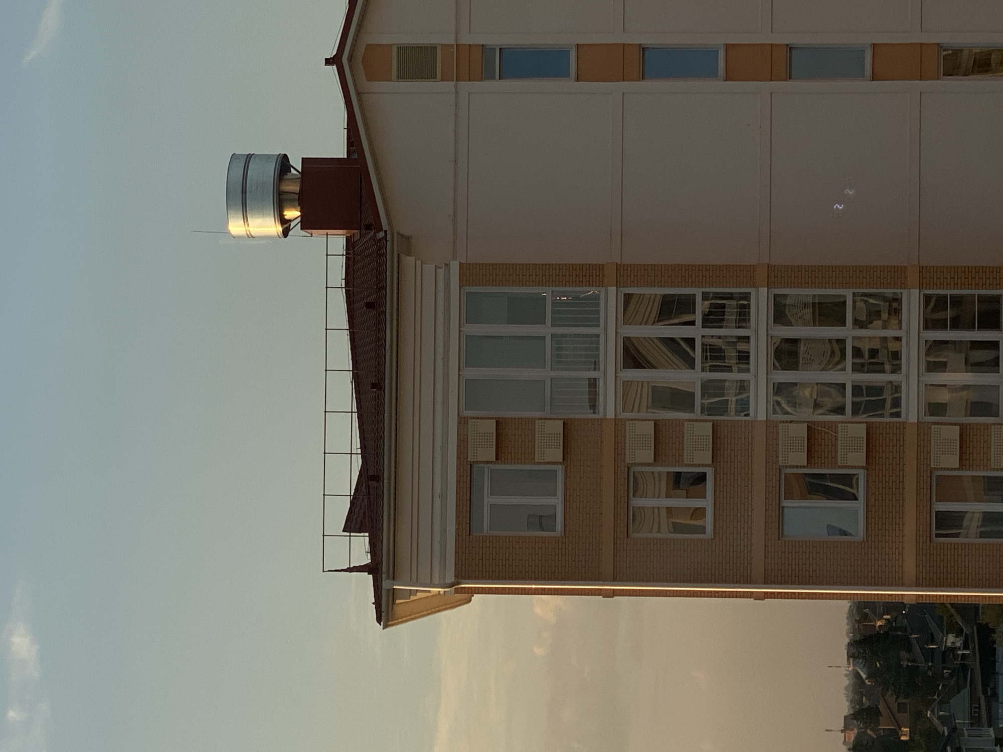 This is a medium shot of a modern apartment building in the city of Stopyatyy, Russia, taken at sunset. The building has a facade of light brown brick on the left and a light cream color on the right. The left side features multiple windows with white frames, some with air conditioning units installed below them. On the right side, there are vertical windows with blue glass, separated by orange trim. The roof is tiled in a reddish-brown color, with a metal railing along the edge. A large, cylindrical metal chimney with reflective bands is prominently positioned on the roof, catching the golden light of the setting sun. The sky above is a gradient of pale blue and orange, with a few wispy clouds. In the distance, a lower-rise urban landscape with trees and rooftops is visible. The image is static and shows no people or animals. The lighting suggests it is late afternoon or early evening.