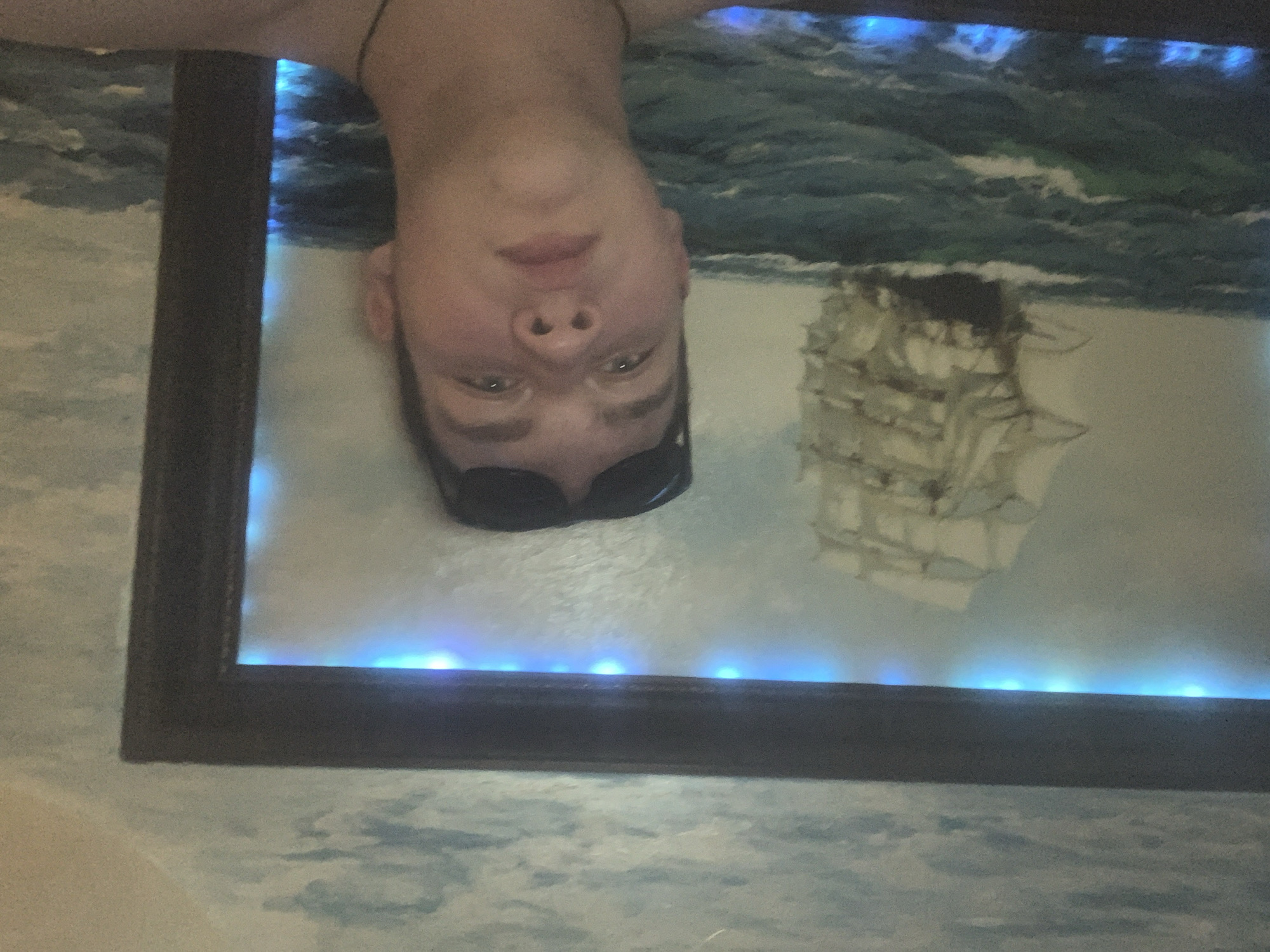 A man is upside down, reflected in what appears to be a framed artwork with a painting of a ship at sea, which has blue LED lights mounted around the inner frame. The background beyond the frame appears to be a similarly themed wallpaper with cloud and sea motifs. The specific location of this scene in Severodvinsk, Russia, cannot be determined from the image alone.